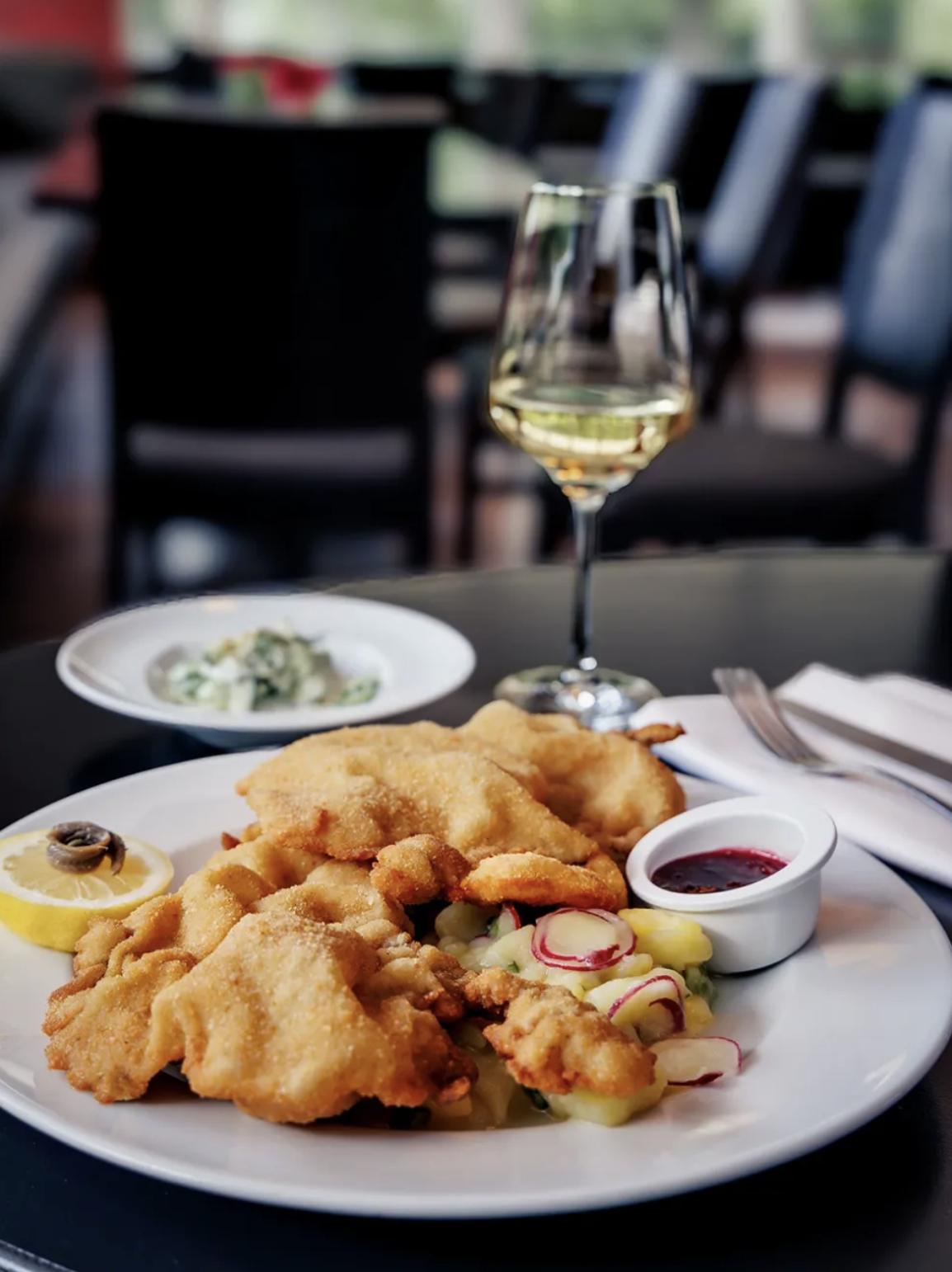 A plate of Schnitzel with lemon, coleslaw, and tartar sauce on a black table, with a glass of white wine and a side dish in the background.