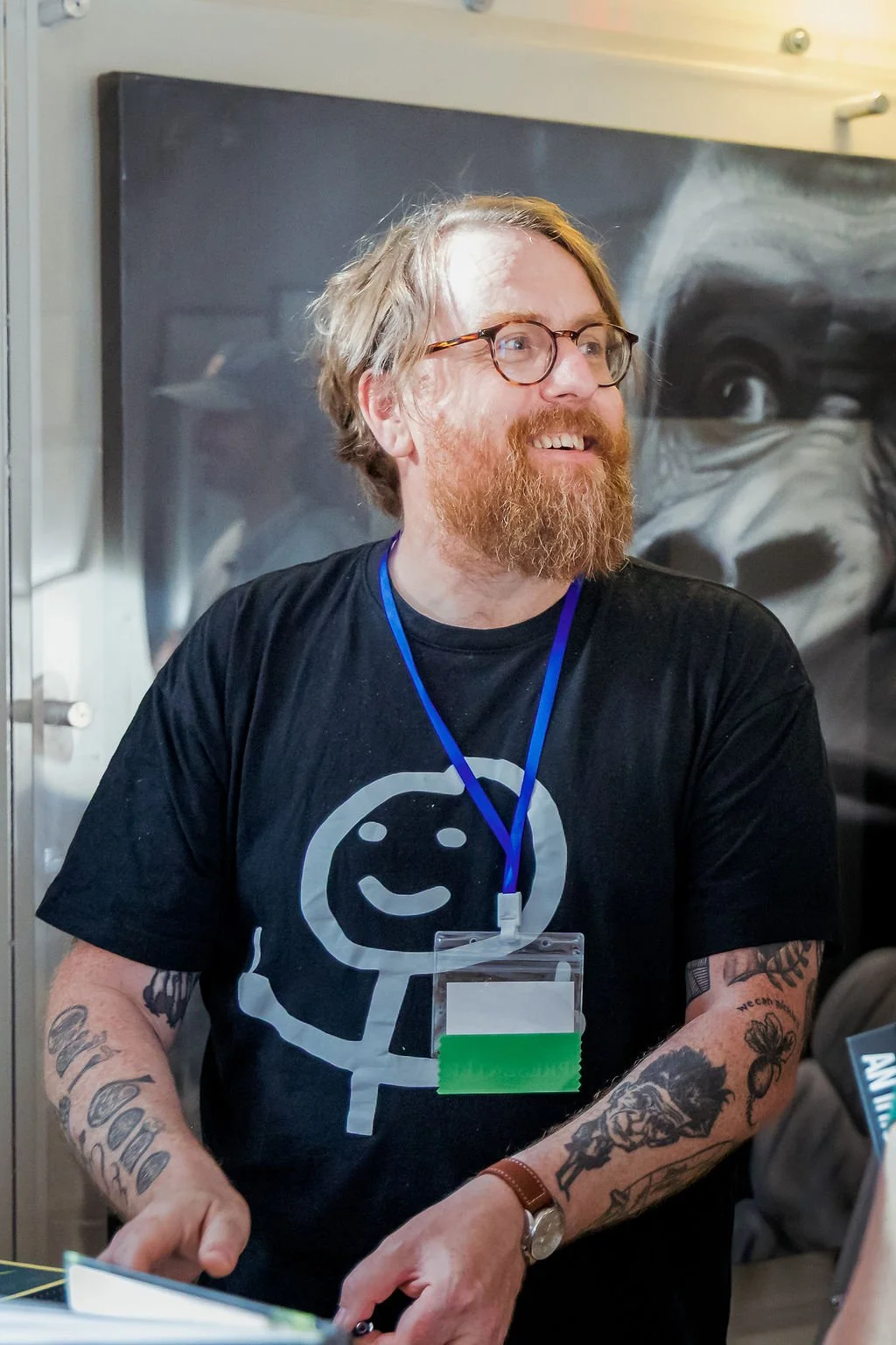 A man with glasses and a beard wearing a black T-shirt with a smiley face logo, standing indoors, smiling, with tattoos on his arms, and wearing a blue lanyard and a watch.