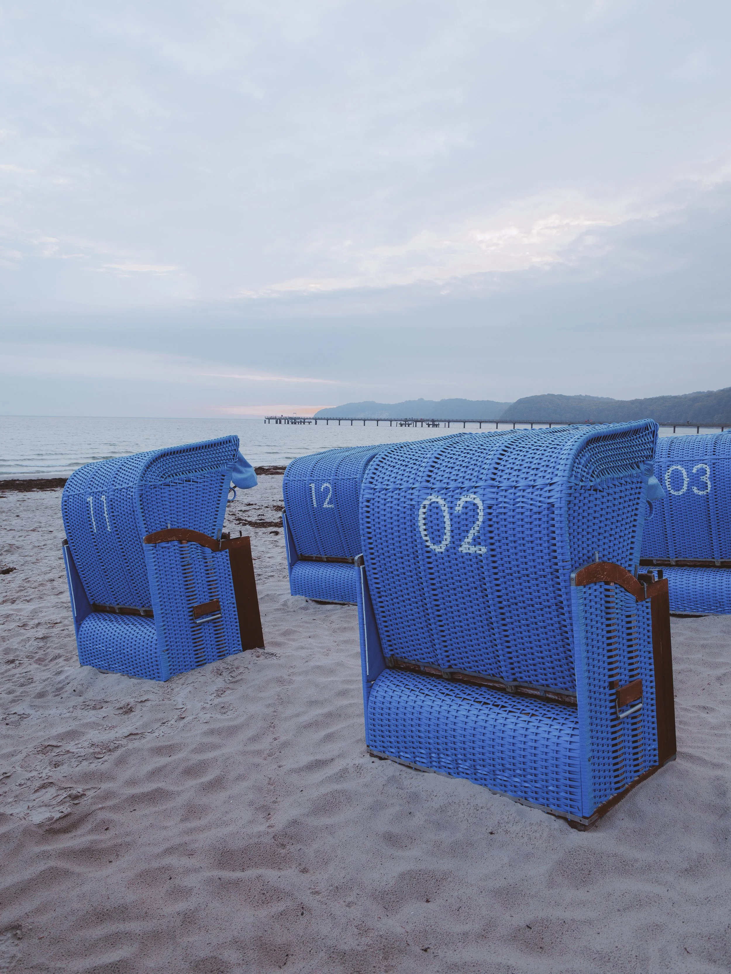Blue woven beach chairs set up on sandy beach with calm ocean and distant pier and hills in the background under cloudy sky.