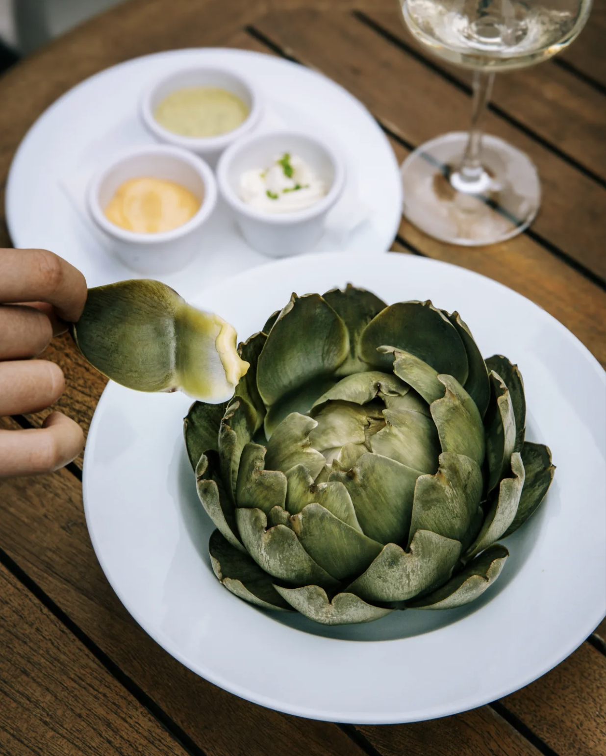 A large roasted artichoke on a white plate, with a hand holding a spiny leaf. In the background, part of a glass of white wine and a white plate with three small bowls containing dips or sauces, set on a wooden table.