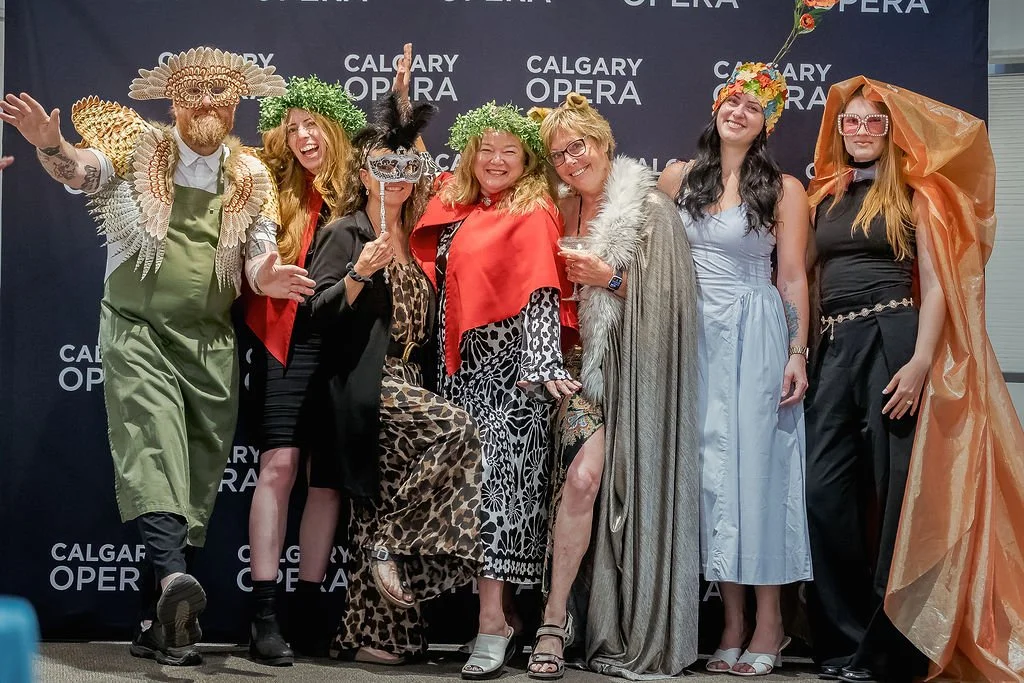 Group of seven people dressed in elaborate costumes, standing in front of a Calgary Opera backdrop, celebrating and posing for the camera.