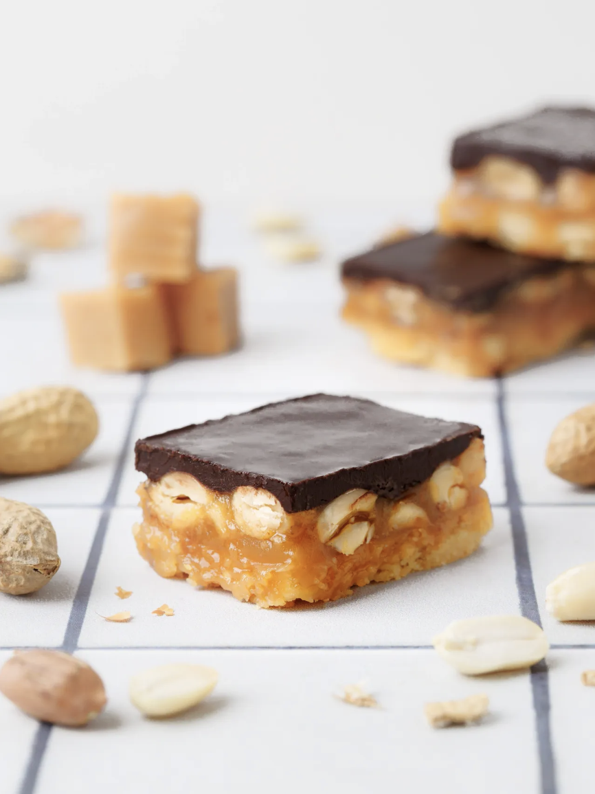 Close-up of a blondie bar with nuts and caramel topped with a chocolate layer, surrounded by other caramel bars and nuts on a white surface.