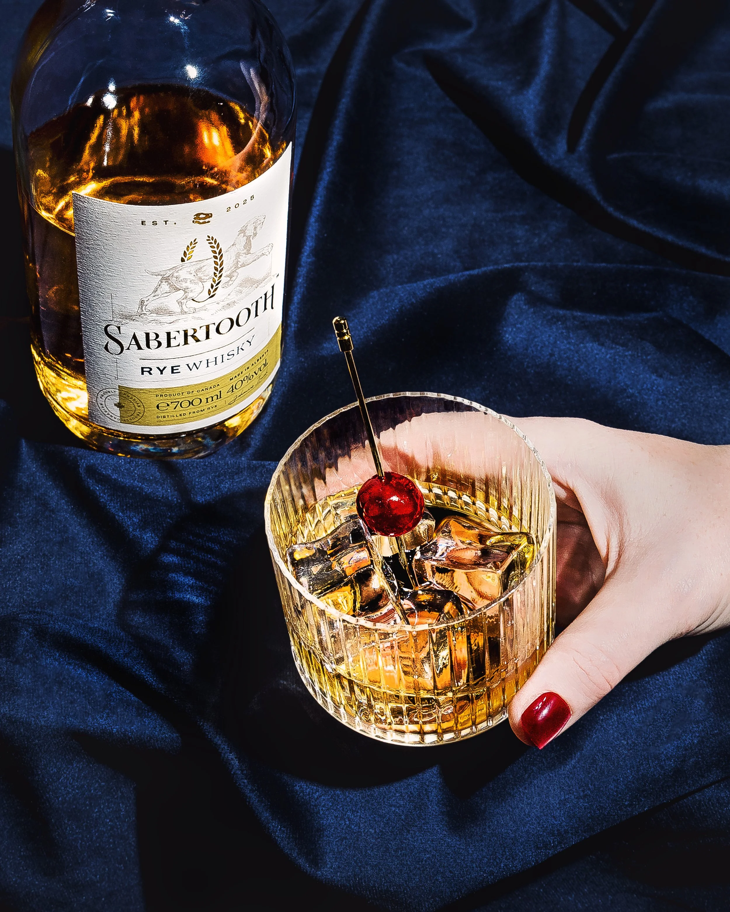 A hand with red nail polish holding a glass of whiskey with ice cubes and a cherry garnish, with a bottle of Sabettooth rye whiskey in the background, on a dark blue satin cloth.