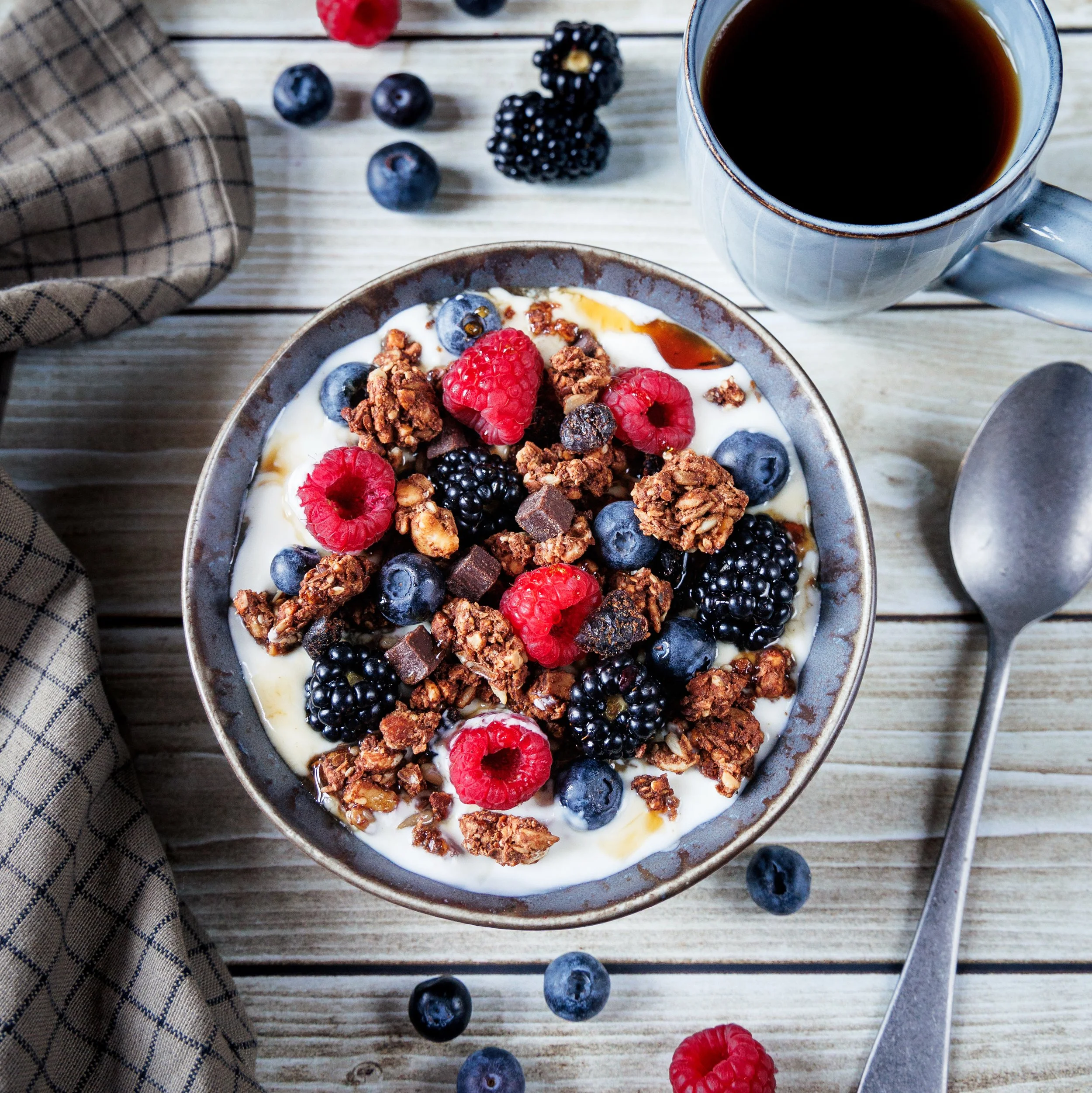 A bowl of yogurt topped with granola and fresh red raspberries and blueberries, with a cup of black coffee nearby on a wooden table. Scattered blueberries and a red raspberry are also on the table beside a checkered cloth.