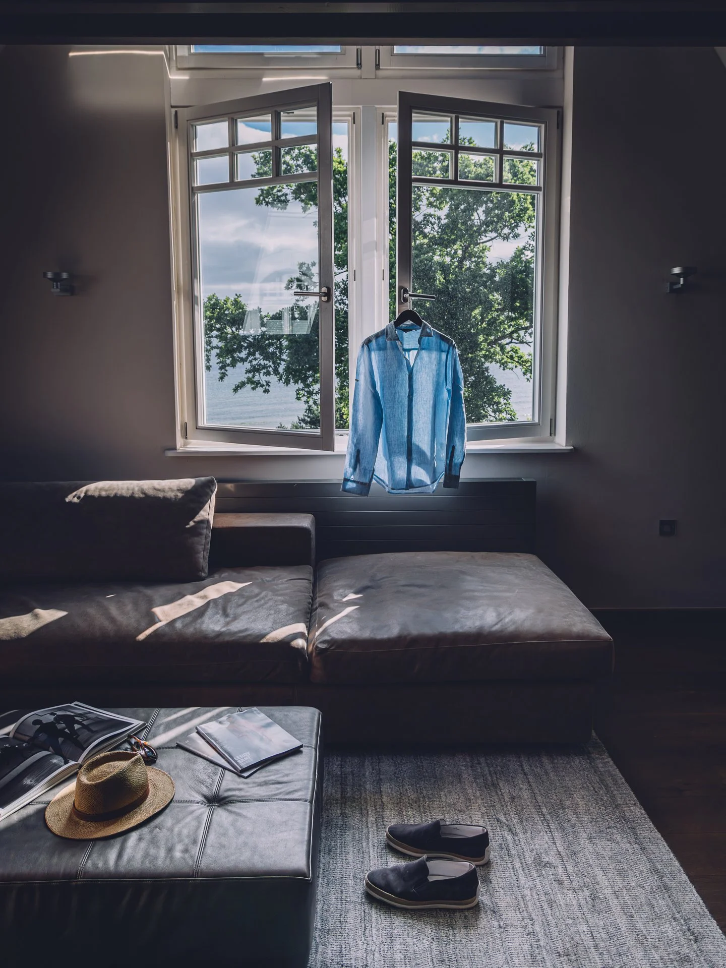 Open window overlooking trees and water, blue button-up shirt hanging on window latch, items on dark leather couch including a straw hat, magazines, and sunglasses, shoes on light-colored rug.