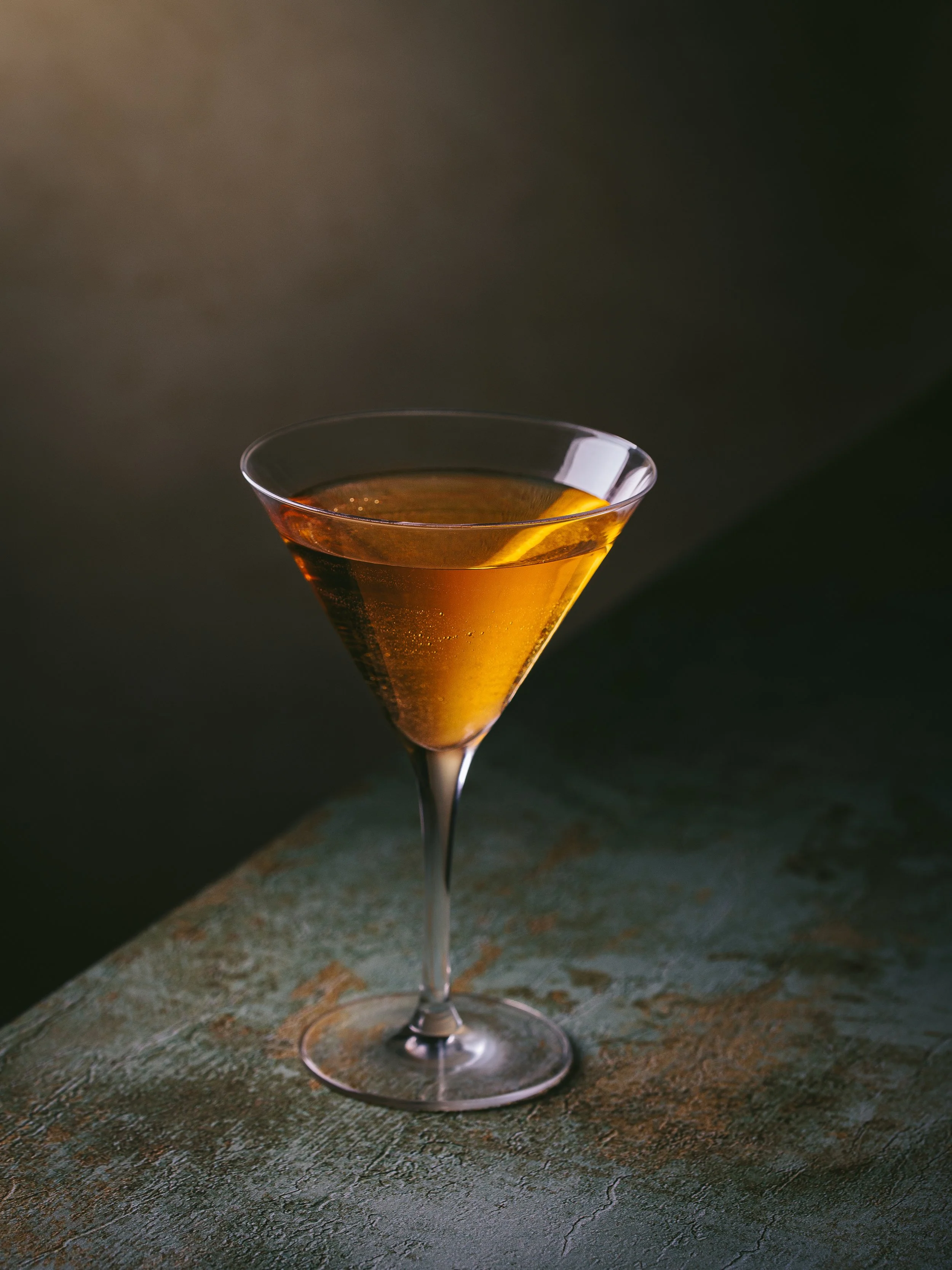 A martini glass filled with an amber-colored cocktail, garnished with a lemon twist, on a textured surface with a dark background.
