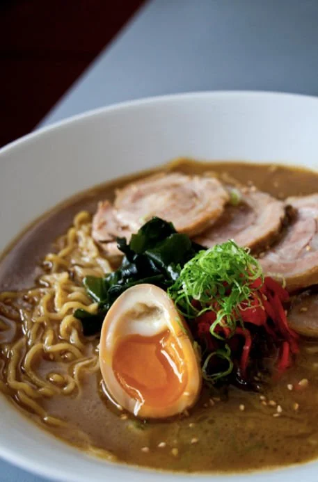 Bowl of ramen with soft-boiled egg, sliced pork, green onions, red pickled ginger, seaweed, and curly noodles in broth.
