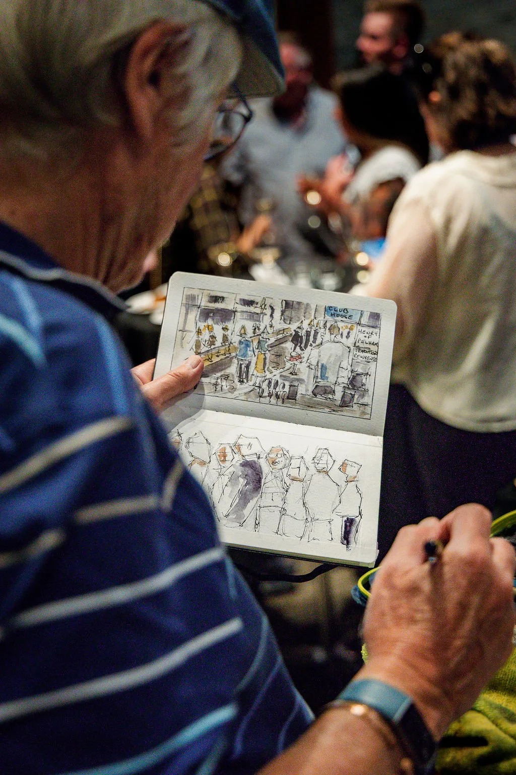 An elderly man with glasses and a striped blue shirt is looking at a sketchbook containing colorful drawings of a busy indoor scene with people and a sign that reads "Club House."