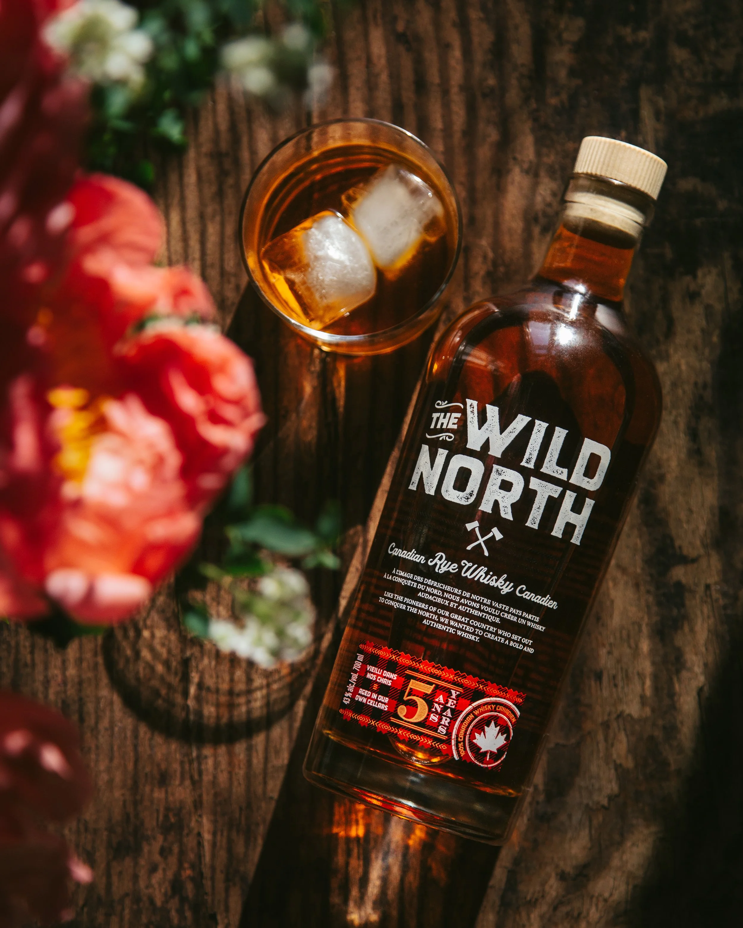 A bottle of Canadian rye whiskey lying on a wooden surface with a glass of whiskey and ice cubes nearby, and some blurred red flowers in the foreground.