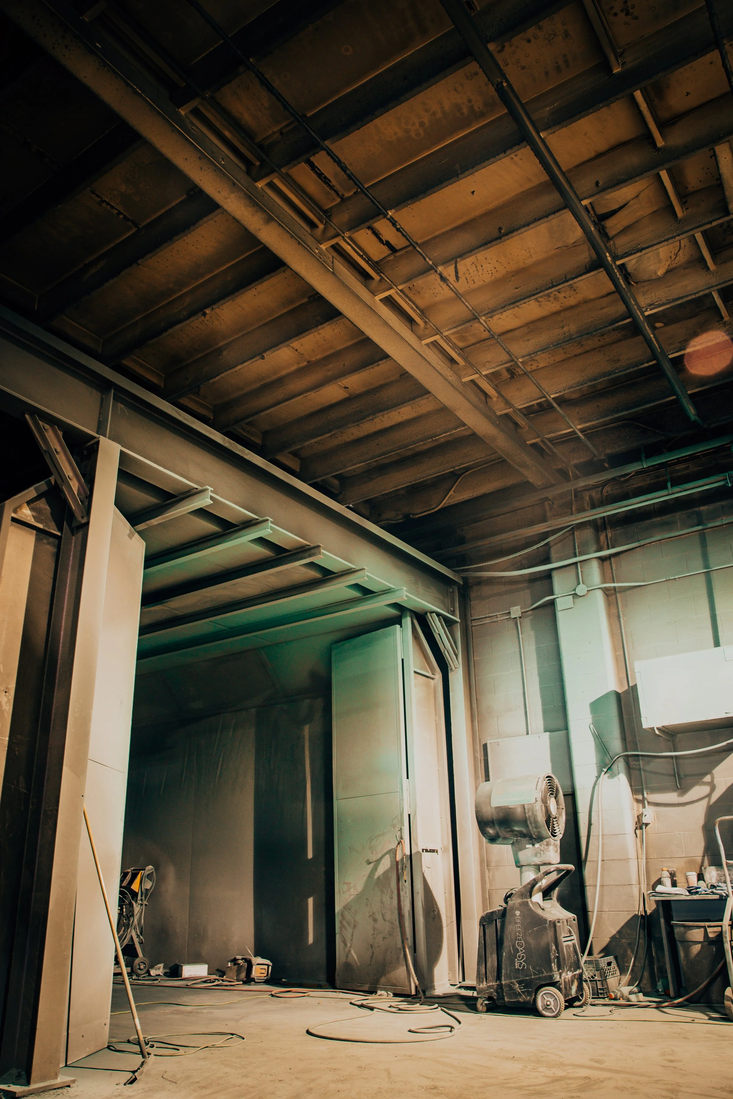 Construction or renovation workspace with tools, equipment, and unfinished walls and ceiling. Includes a metal cabinet or enclosure, a black industrial fan, and scattered electrical cords and tools on the floor.
