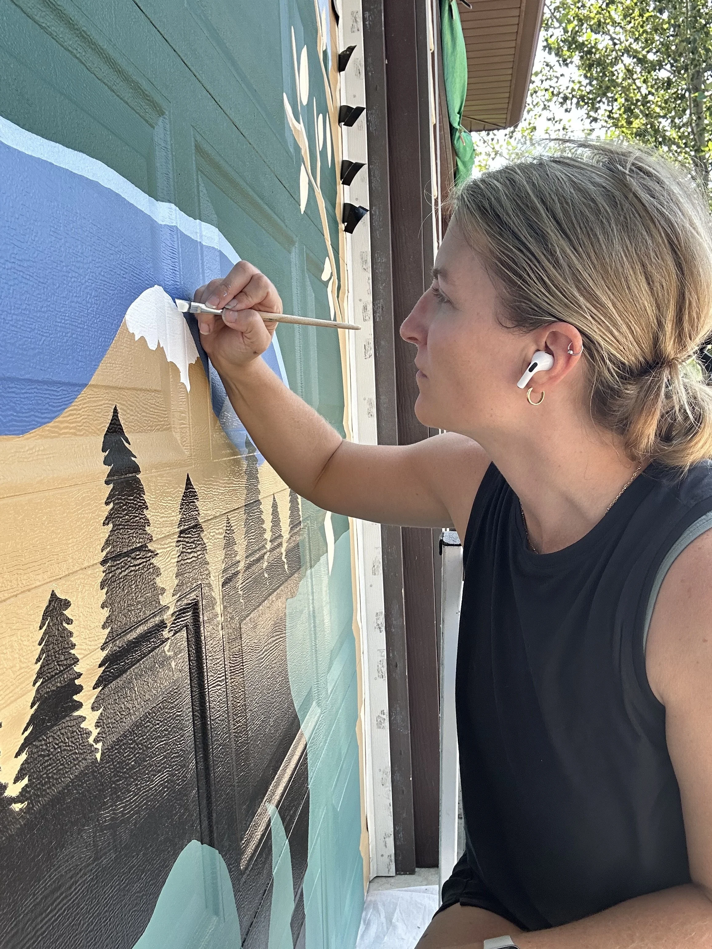 A person with light-colored hair, wearing a black sleeveless shirt, and white wireless earbuds, painting a mural depicting a mountain landscape, trees, and a lake on a wooden wall outdoors.