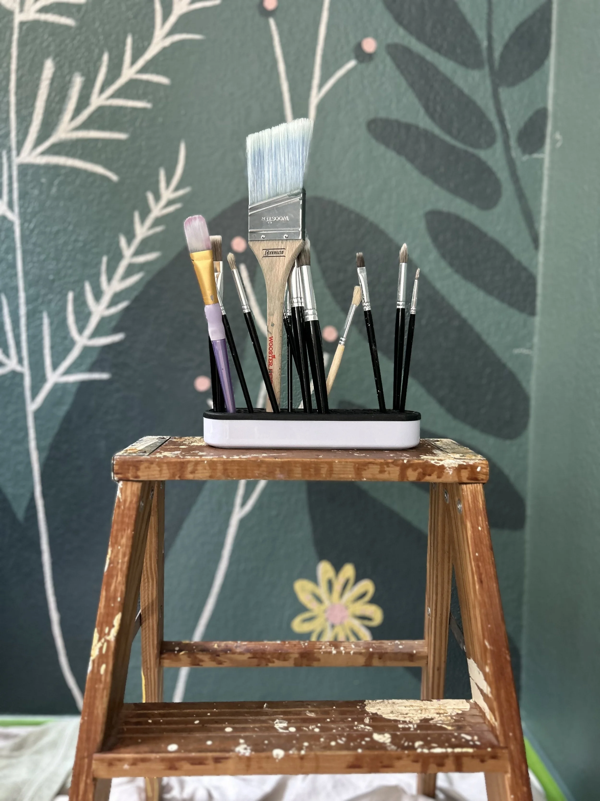 A wooden step ladder with paint splatters, holding various paintbrushes, standing in front of a painted wall with botanical designs including leaves and flowers.
