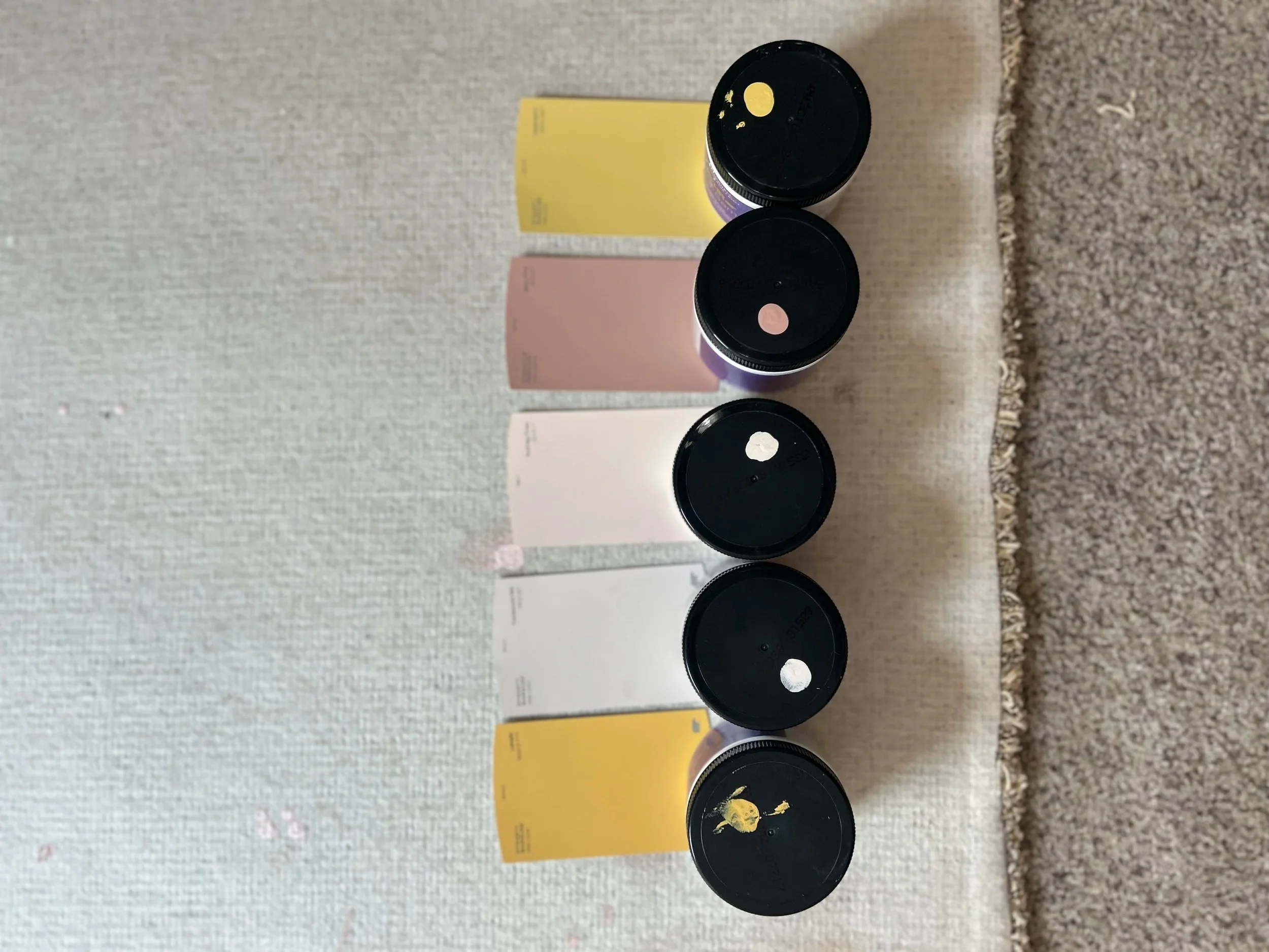 Four black jars with black lids, two of which have paint splatters on top, are placed on a beige textured surface. Behind the jars, there are colorful rectangular cards in yellow, pink, and white, with some text on them, possibly product labels or packaging.