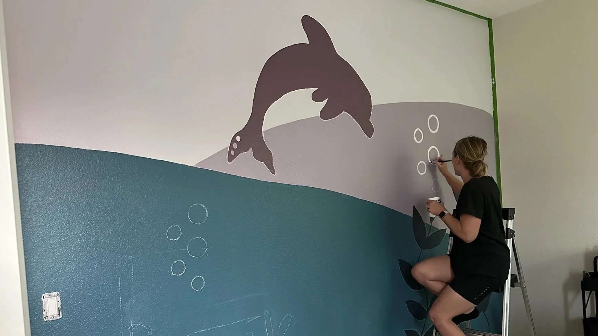 A woman is painting a mural on a wall, depicting a dolphin jumping out of water with bubbles around it. The wall has a blue and gray background with minimalistic scenery.