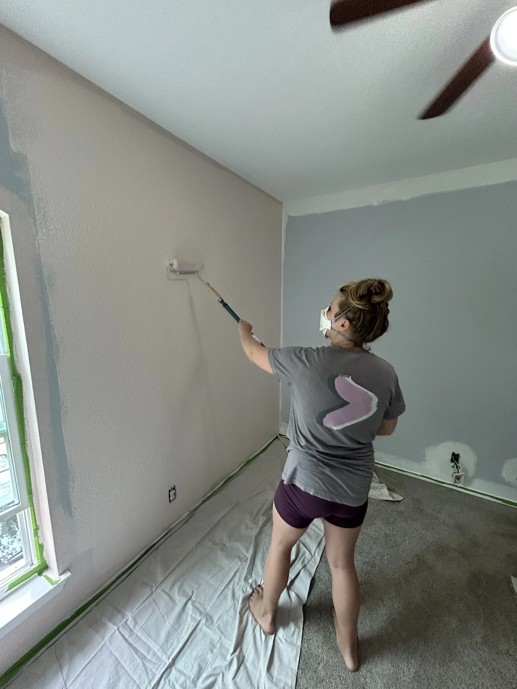 A woman wearing a face mask painting a wall beige with a roller brush inside a room with a window, green painter's tape, and a plastic sheet covering the floor.