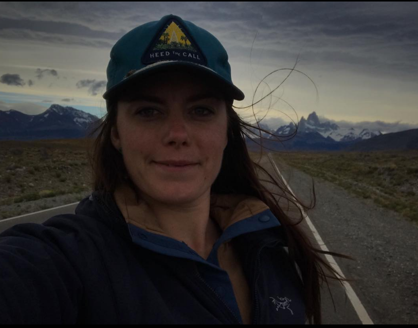 A woman taking a selfie on an empty road with mountains and a cloudy sky in the background, wearing a blue cap and a jacket.