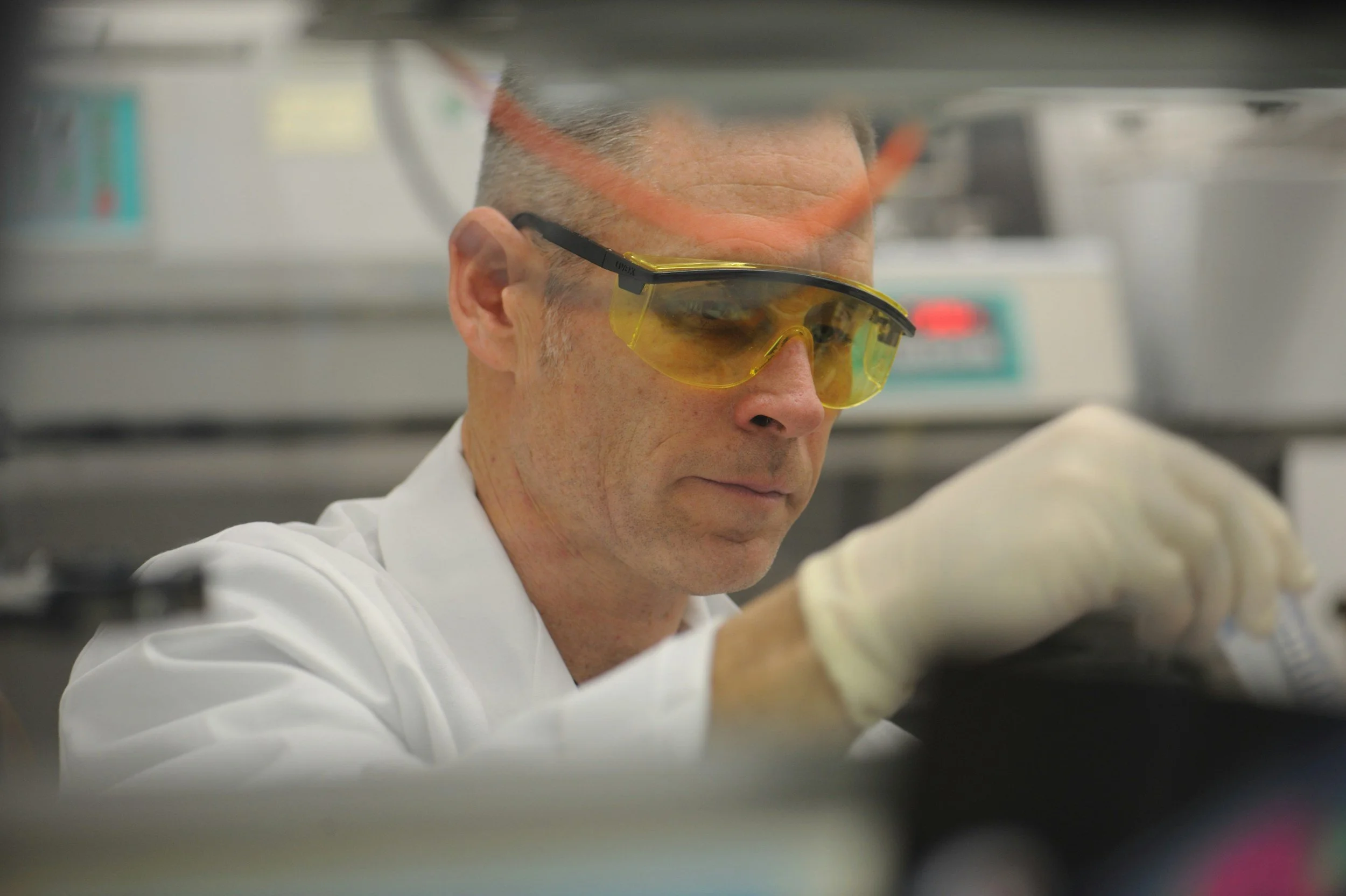 A scientist wearing yellow safety glasses and a white lab coat is working in a laboratory, examining a sample.