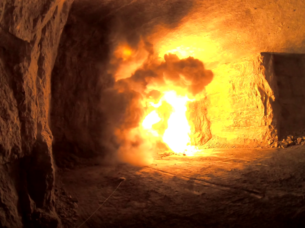 A large fireball and thick smoke inside a tunnel or cave with rocky walls, illuminated by the flames.
