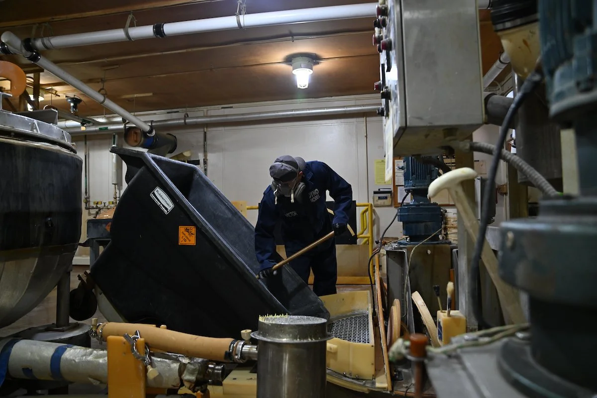A worker in protective gear scooping a substance from a black container in an industrial setting with pipes and machinery.
