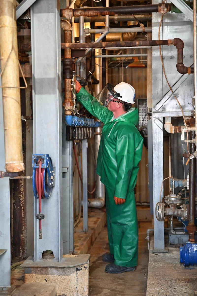 A worker in green rain gear and a white helmet inspecting pipes in an industrial setting.