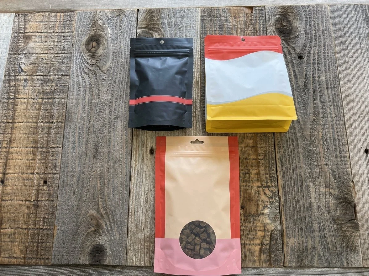 Three resealable stand-up pouches on a rustic wooden surface, including black, yellow-and-white, and pink packaging; the bottom pouch has a clear window showing small brown treats.