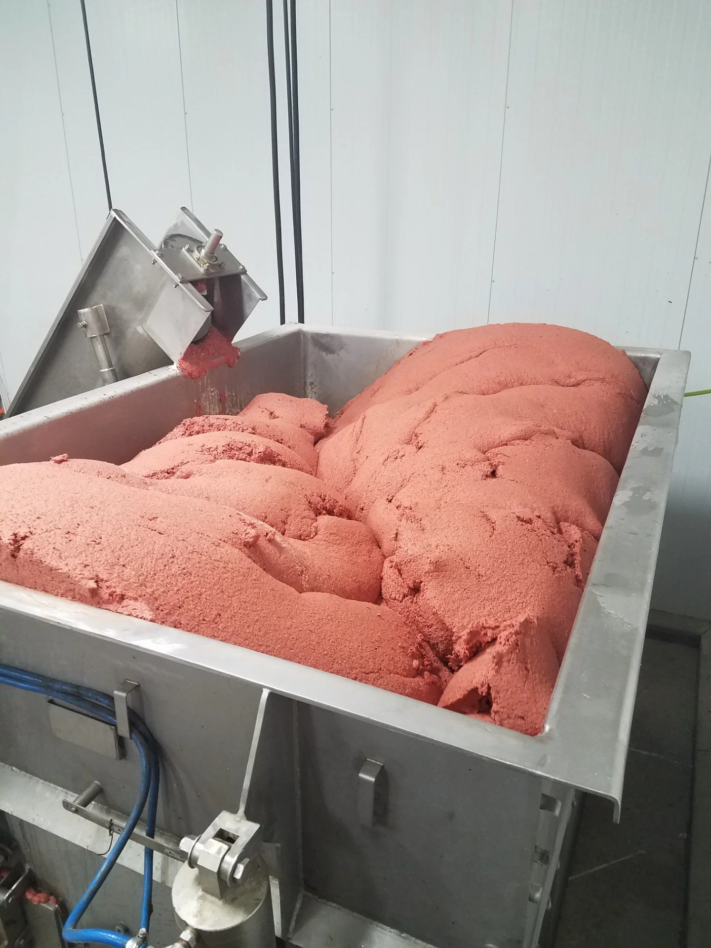 Industrial stainless steel mixing tank filled with pink processed food mixture and a metal hopper dispensing more material.