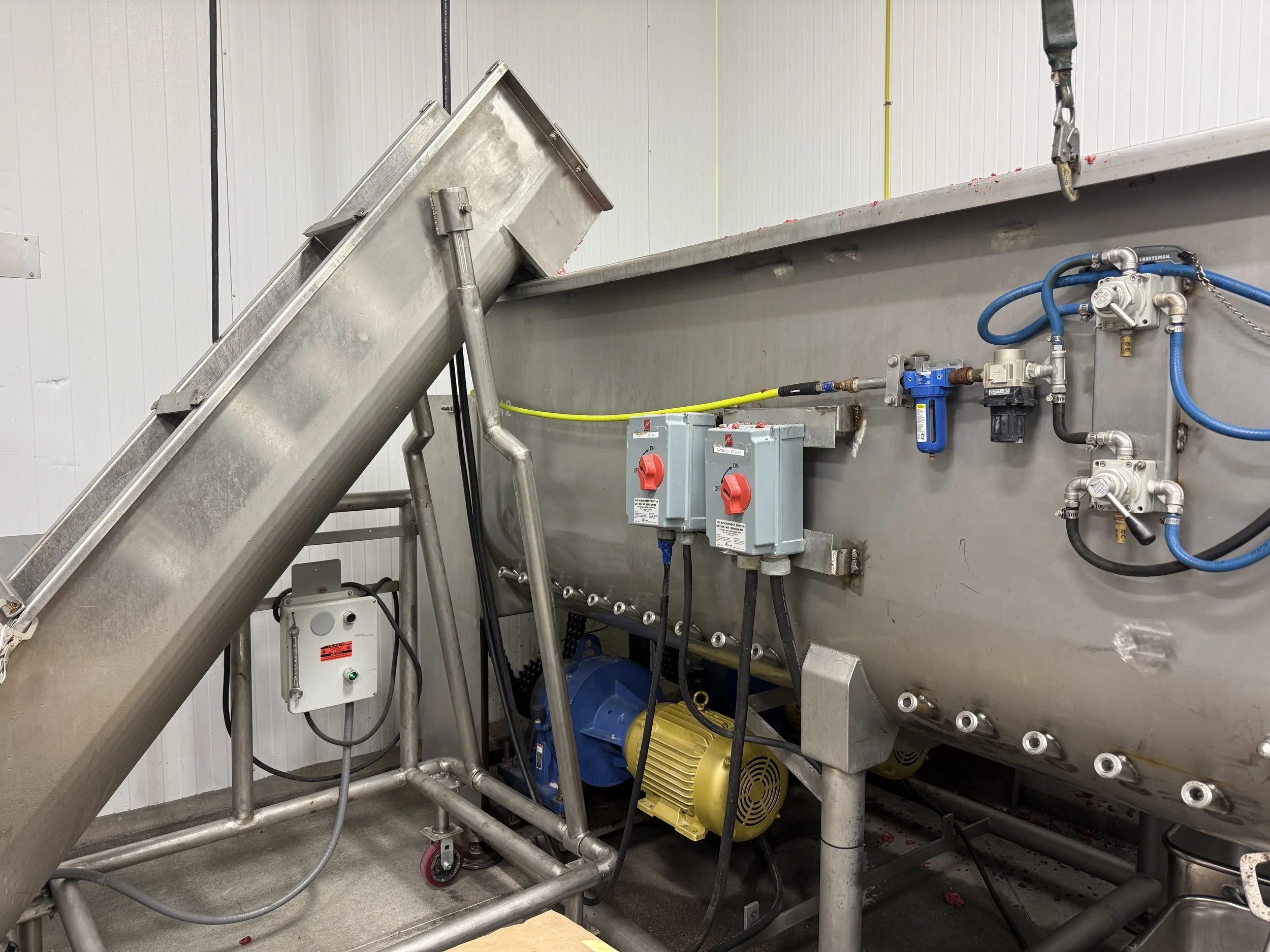 Industrial stainless steel processing tank with inclined hopper, pumps, electrical controls, and blue pneumatic hoses in a food processing facility