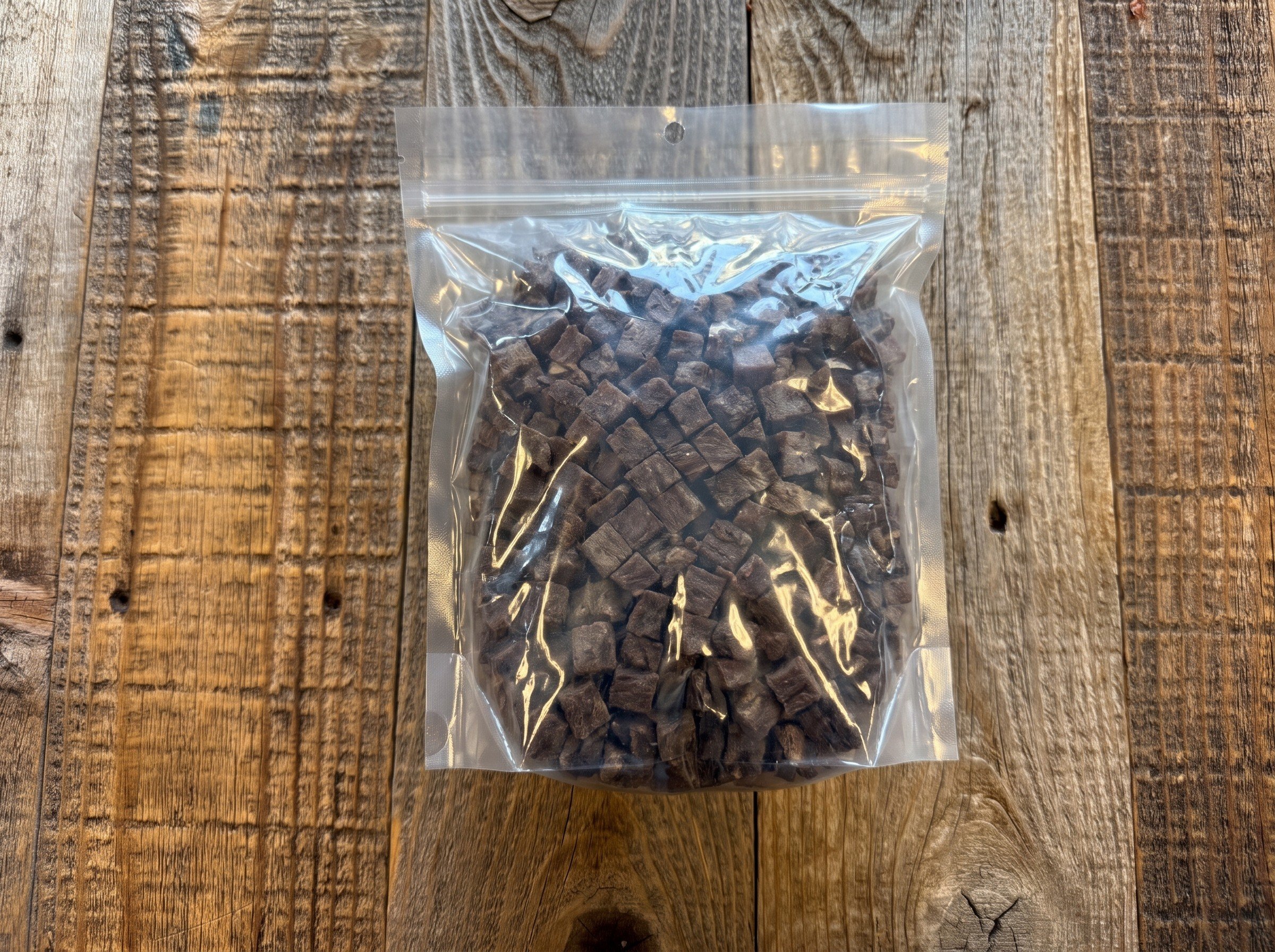 Clear resealable bag filled with small dark brown beef treats cubes on a rustic wooden surface