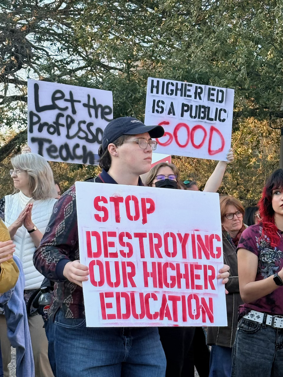 Group of people protesting with signs, including one that reads 'STOP DESTROYING OUR HIGHER EDUCATION' and others advocating for teachers and public education.