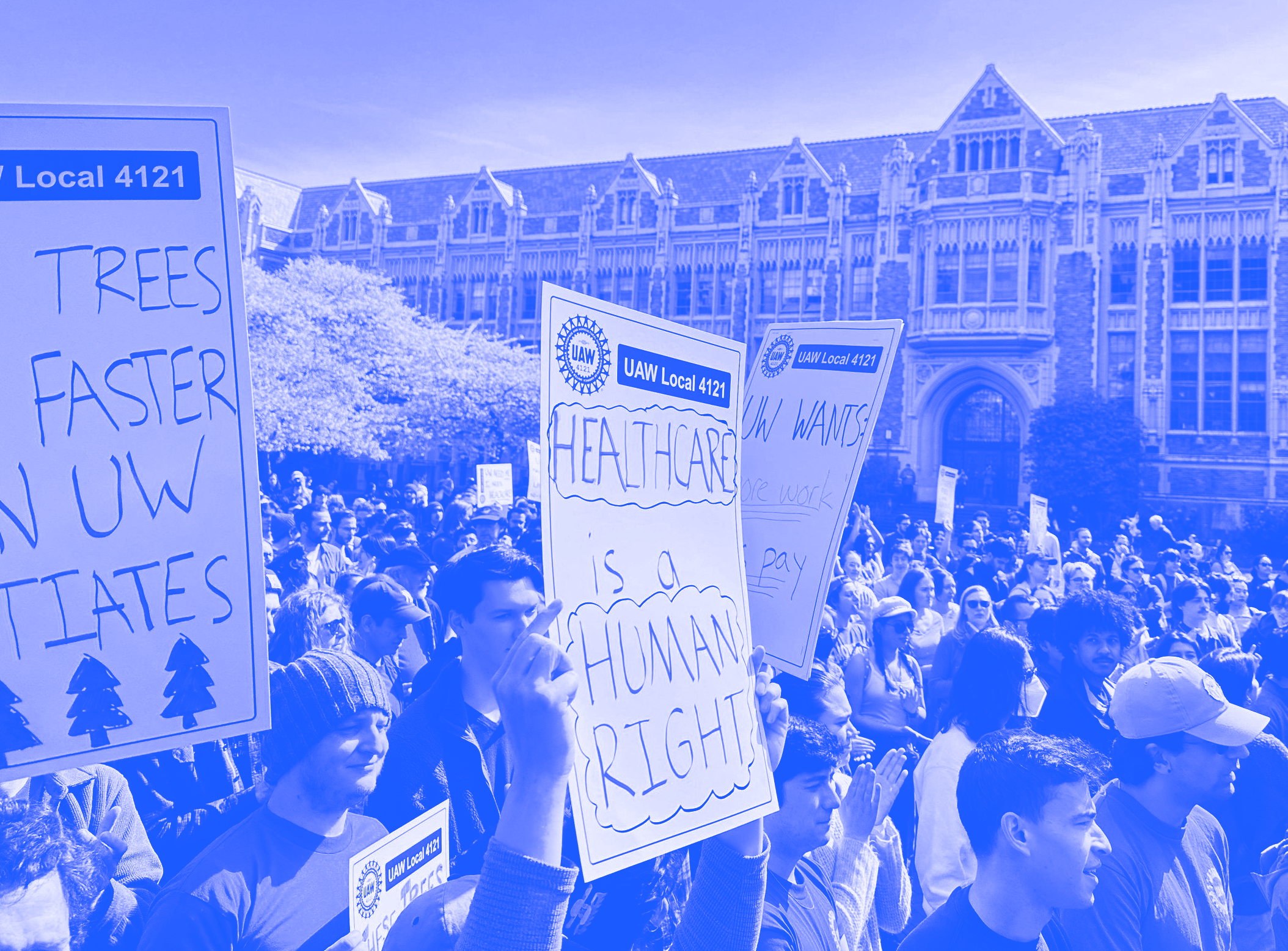 Crowd of people holding protest signs in front of a historic building with ornate architecture. Some signs include messages about trees, healthcare, and wages.