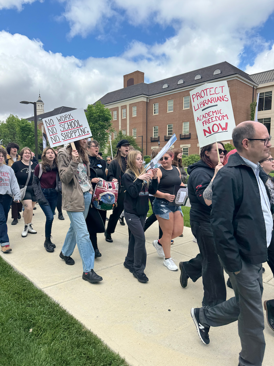 A group of people participating in a protest or rally, holding signs with messages about protecting librarians and academic freedom, walking on a sidewalk near a brick building with a cloudy sky overhead.