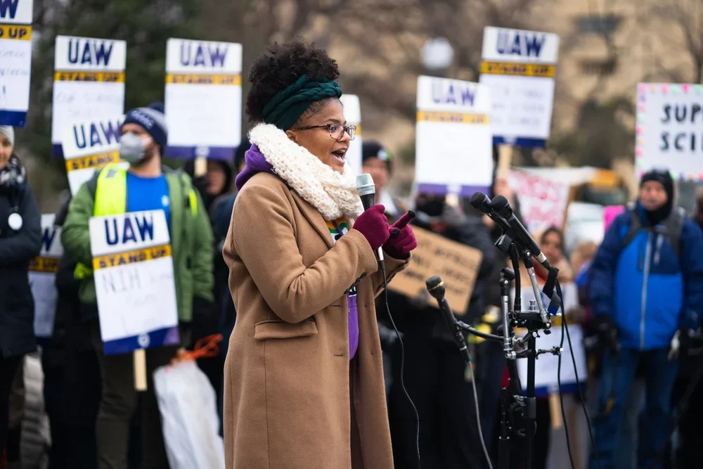 A woman speaking into a microphone at a protest or rally, with people holding signs behind her.