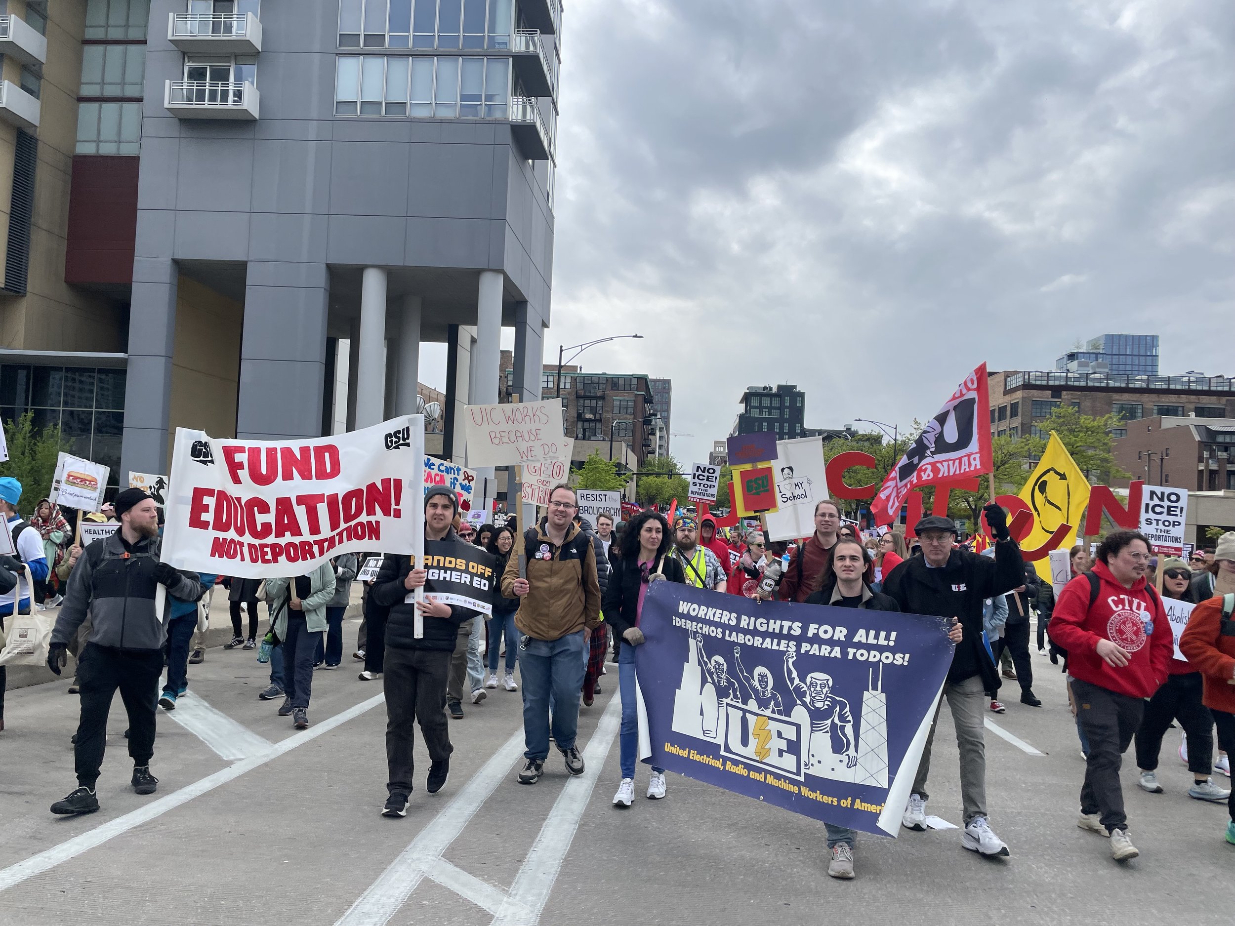 A large crowd participating in a protest march on an urban street, holding various signs and banners advocating for workers' rights and education funding, with tall buildings in the background under a cloudy sky.