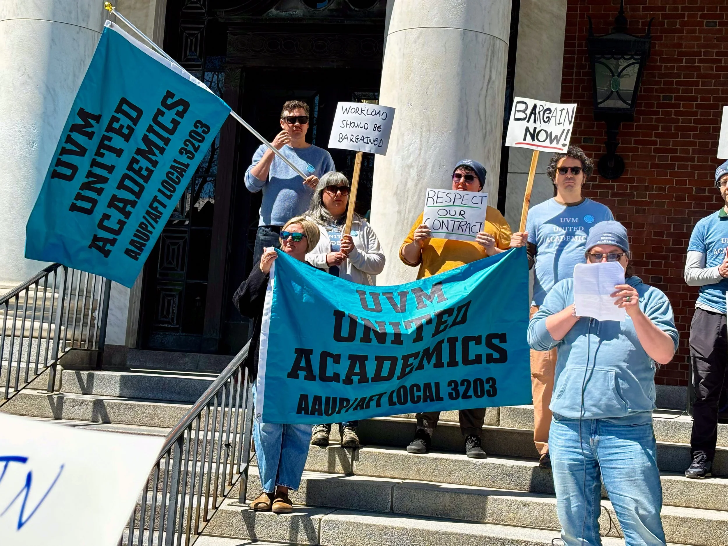 Group of protesters standing on the steps of a building. They are holding signs and banners . One banner reads 'UVM United Academics.'