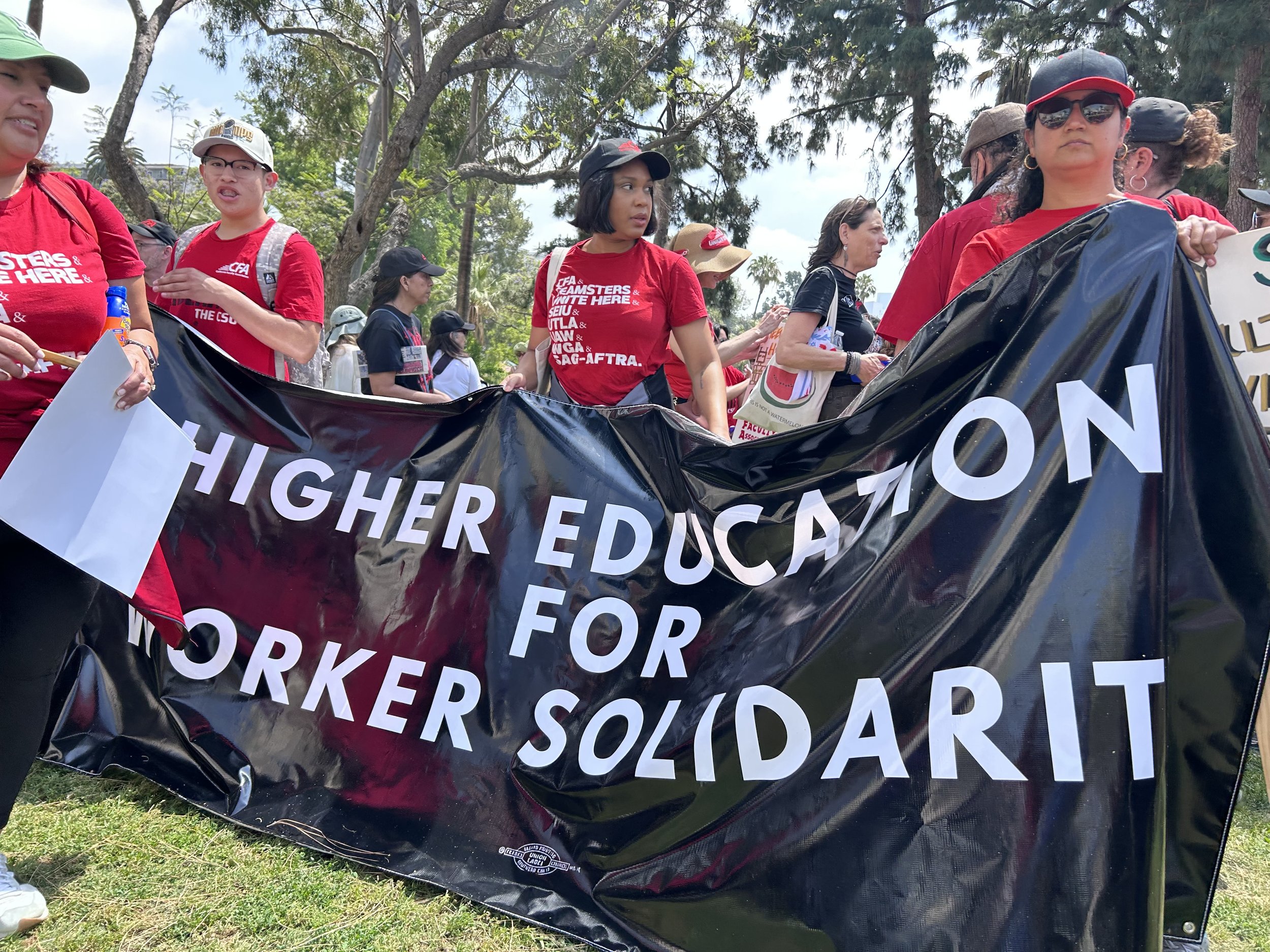 Group of people participating in a protest or rally outdoors, holding a large black banner with white text, surrounded by trees and clear sky.