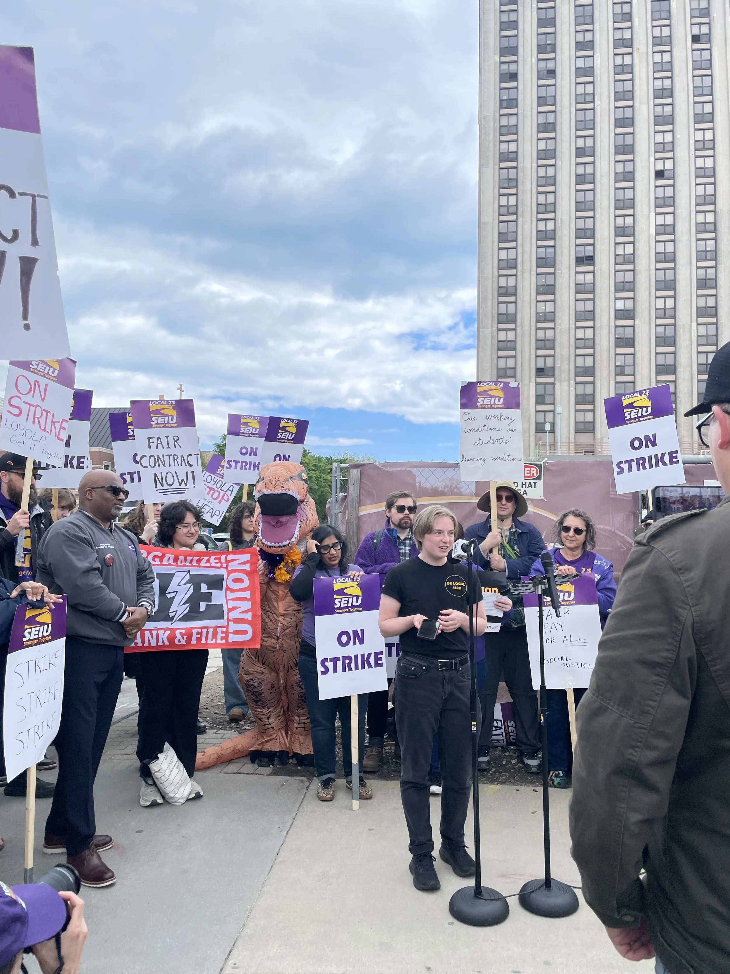 People protesting on a city street, holding signs supporting a strike and demanding fair contracts. Some signs read 'ON STRIKE!' and 'FAIR CONTRACT NOW!' in front of a large building with a grid of windows.