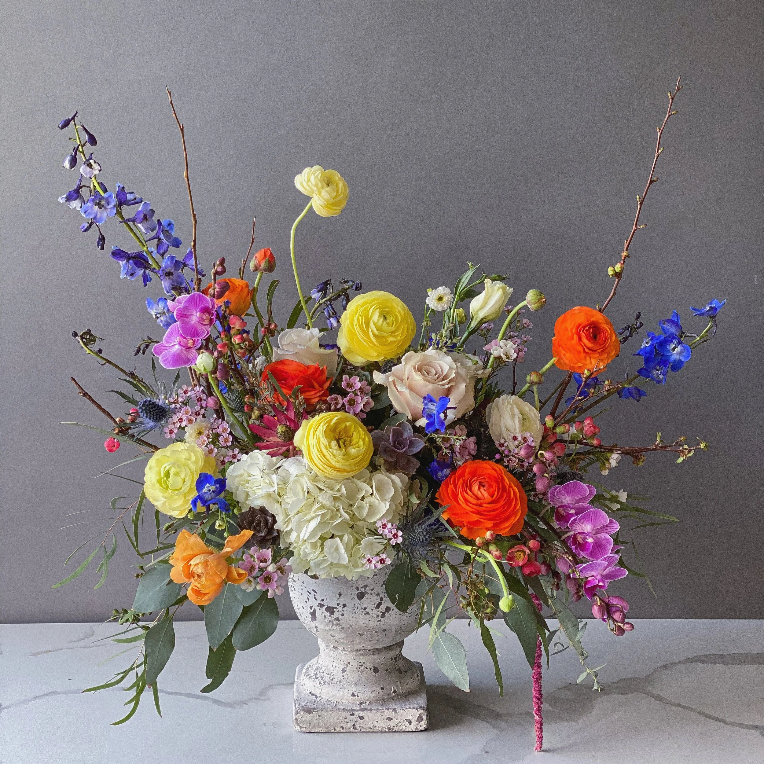 A colorful flower arrangement in a stone vase, featuring purple, yellow, white, orange, and pink flowers along with green leaves and long branches, set against a plain gray background.