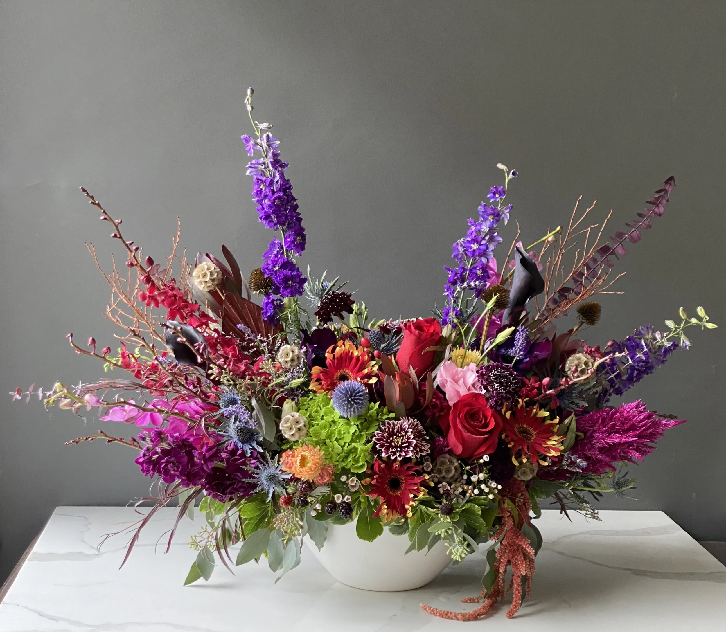 A large colorful flower arrangement in a white vase on a white surface against a gray background.