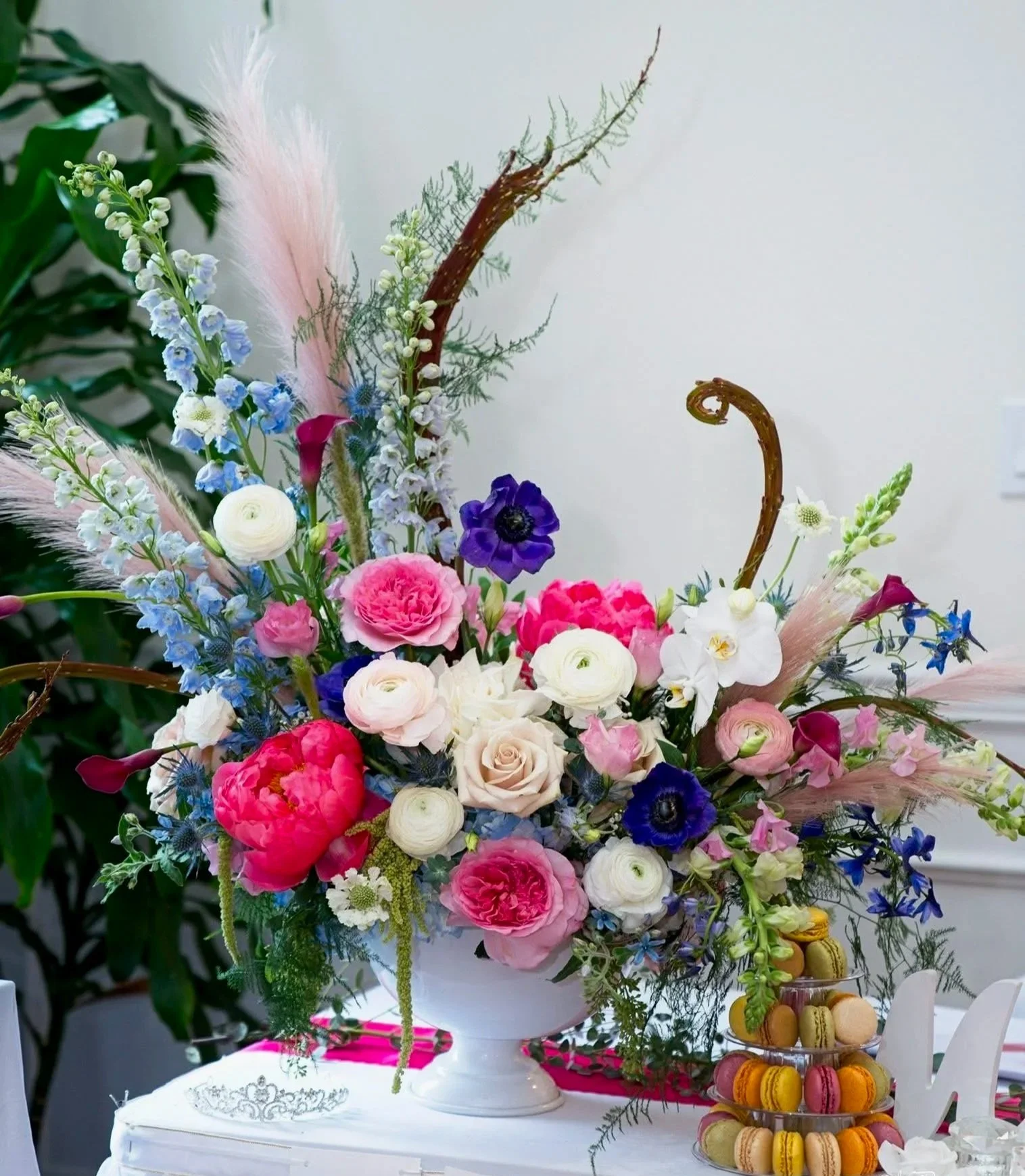 A colorful flower arrangement in a white vase on a table, with a tiered stand of macarons beside it. The flowers include pink, white, purple, and blue blossoms with green foliage and some tall grasses or decorative branches.
