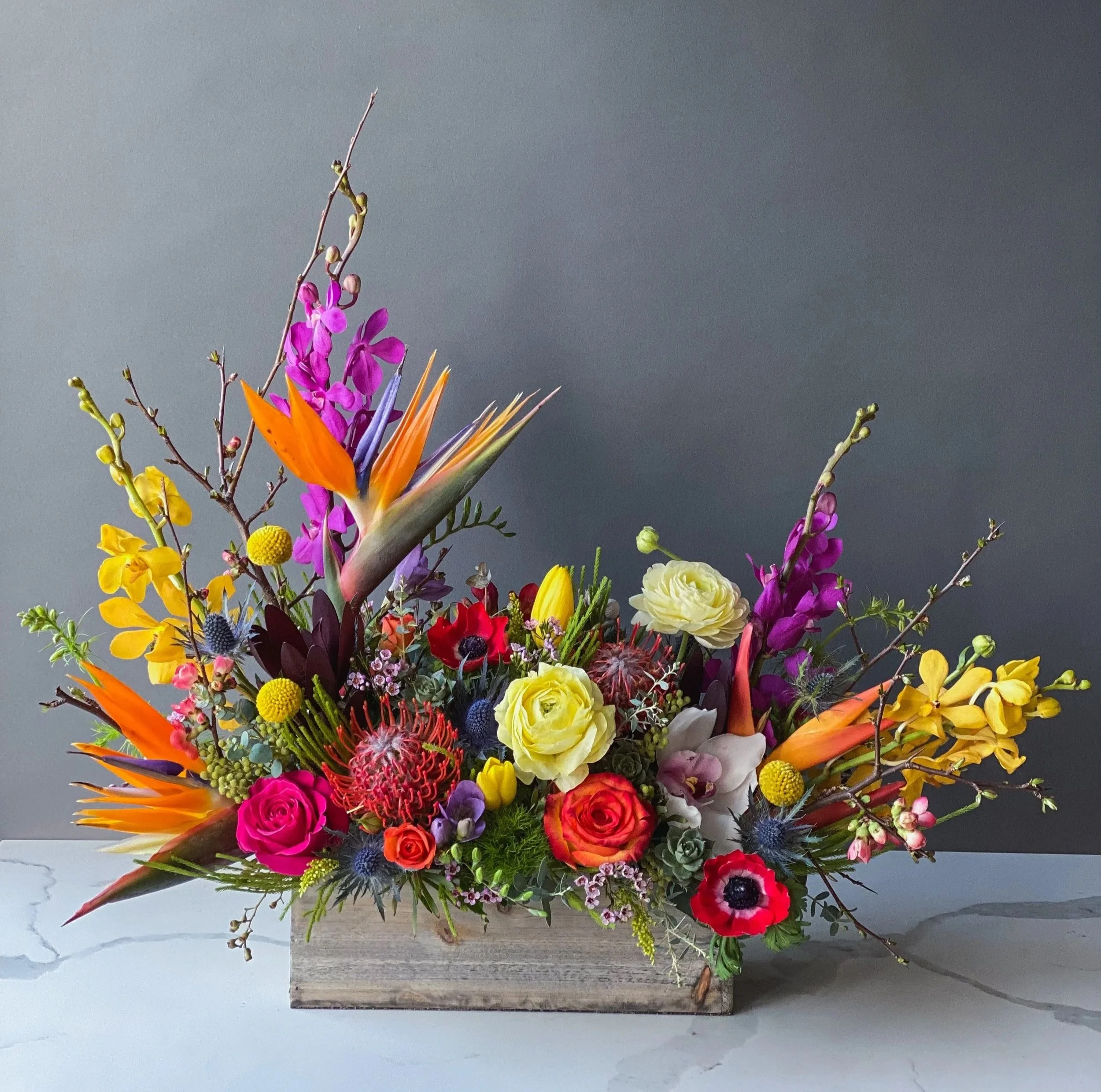 Colorful flower bouquet with pink, yellow, orange, purple, and white flowers arranged in a wooden box against a gray wall.