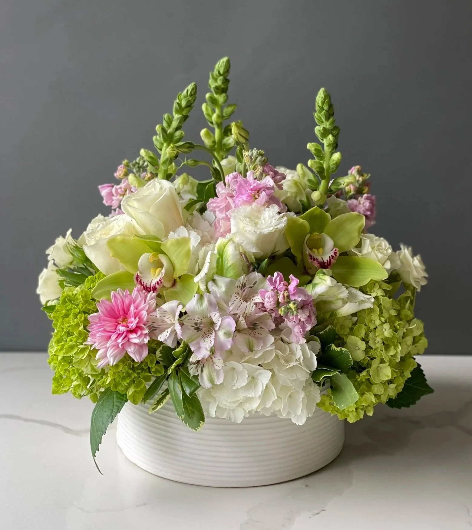 A floral arrangement with white, pink, and green flowers in a white ribbed ceramic vase on a gray surface against a gray background.
