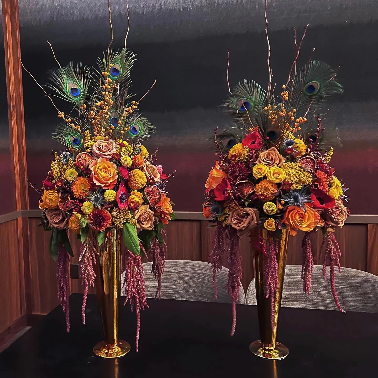 Two large, ornate flower arrangements in gold vases with peacock feathers, yellow berries, roses, and various colorful flowers, set against a dark background.