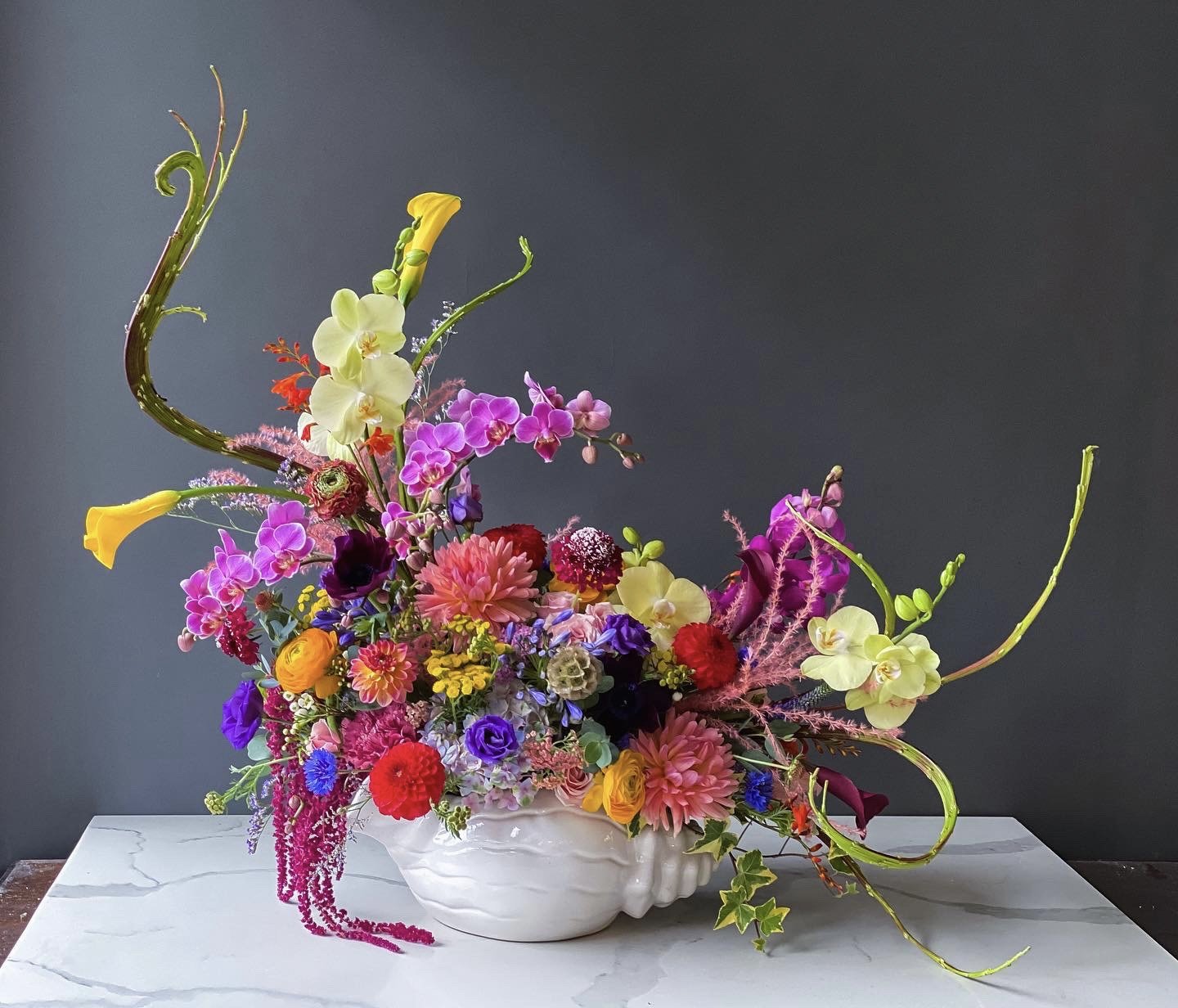 Colorful flower arrangement with various flowers including orchids, roses, dahlias, and greenery in a white ceramic vase on a white marble table against a dark gray background.