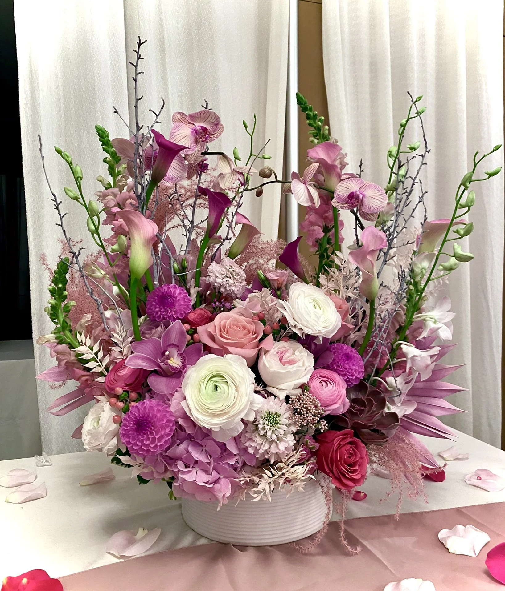 A large floral arrangement with pink, purple, and white flowers in a white round vase on a table decorated with scattered flower petals.