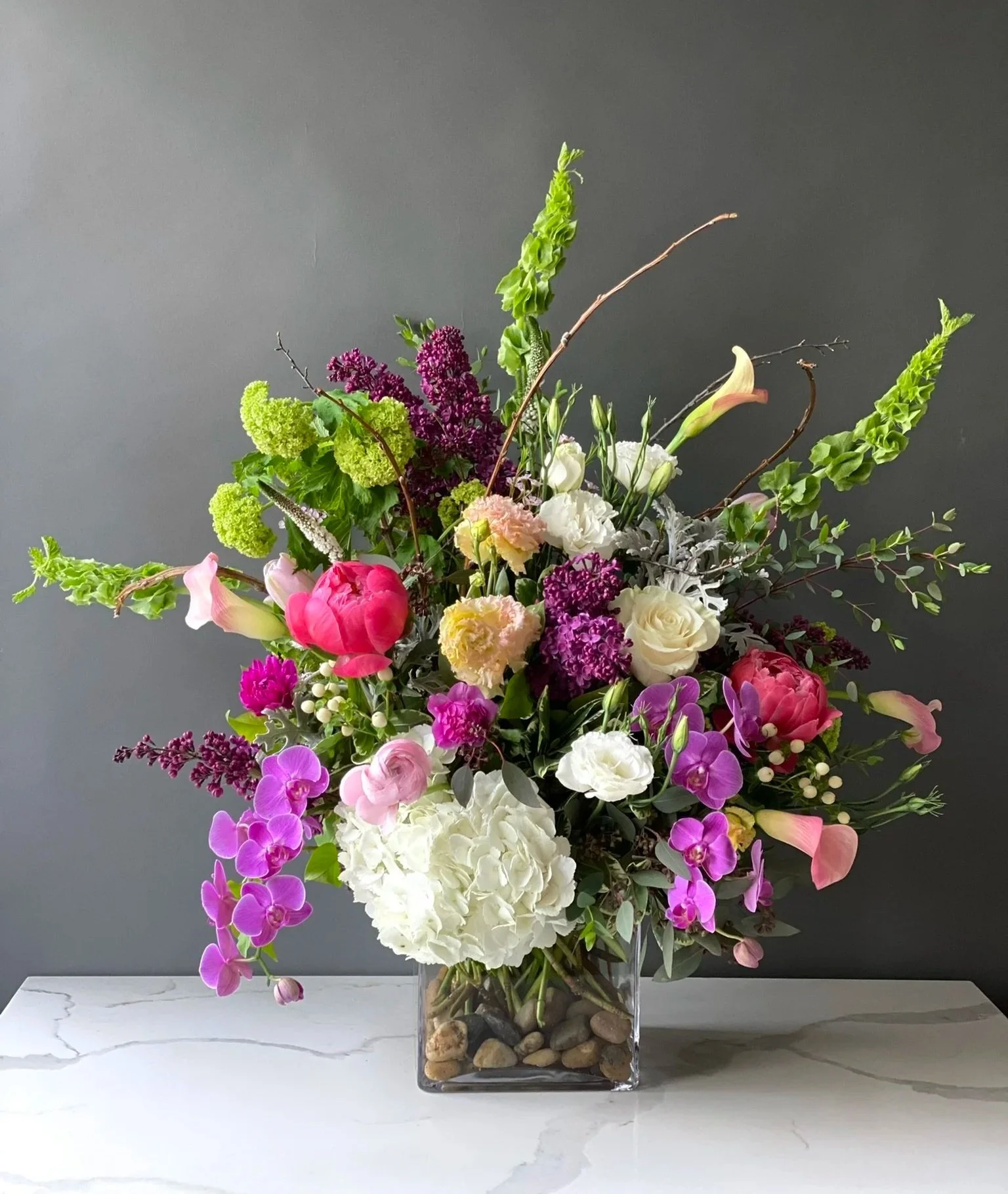 A colorful floral arrangement in a rectangular glass vase filled with rocks, including pink tulips, purple orchids, white roses, pink carnations, and green foliage on a marble surface with a gray background.