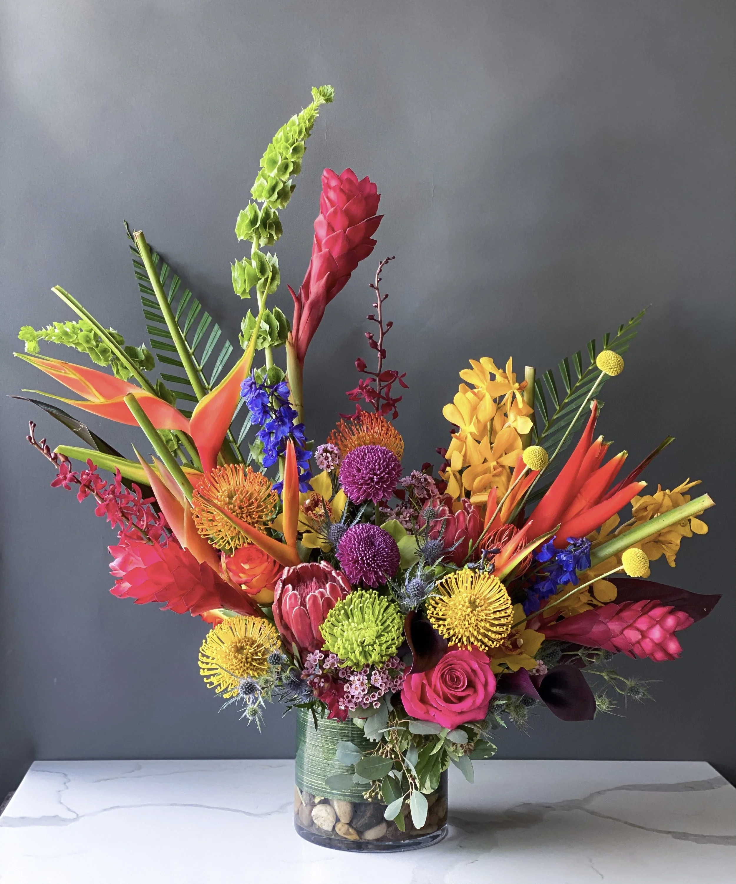 A vibrant multicolored flower bouquet in a glass vase with rocks at the bottom, set on a white marble surface against a dark gray background.