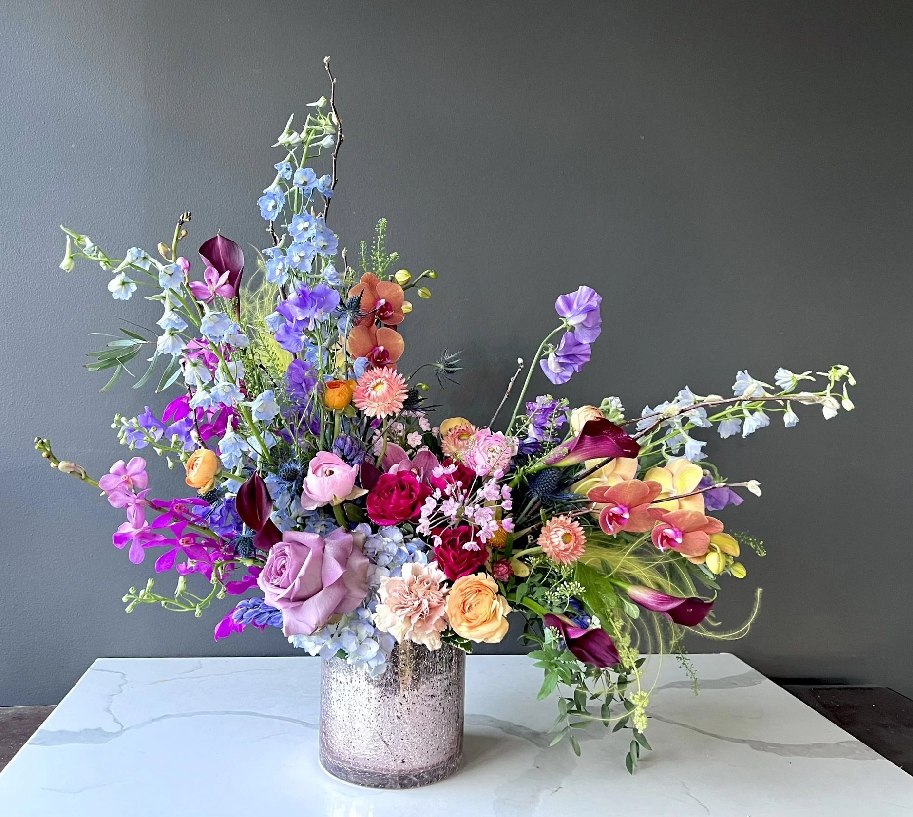 Colorful flower arrangement in a pink speckled vase on a white tabletop against a gray wall background.