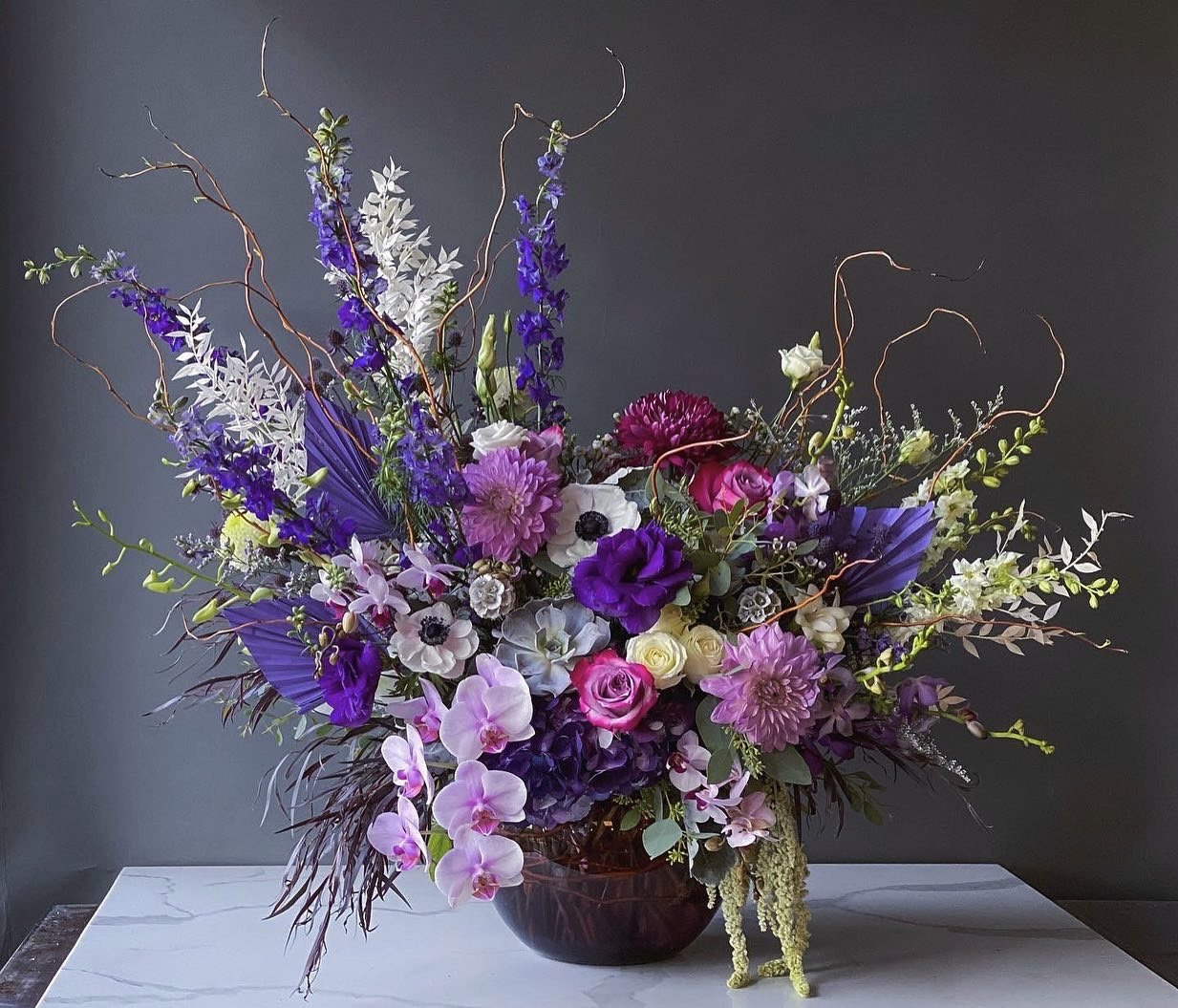 A large, colorful floral arrangement with purple, pink, white, and green flowers in a dark vase on a white marble surface.