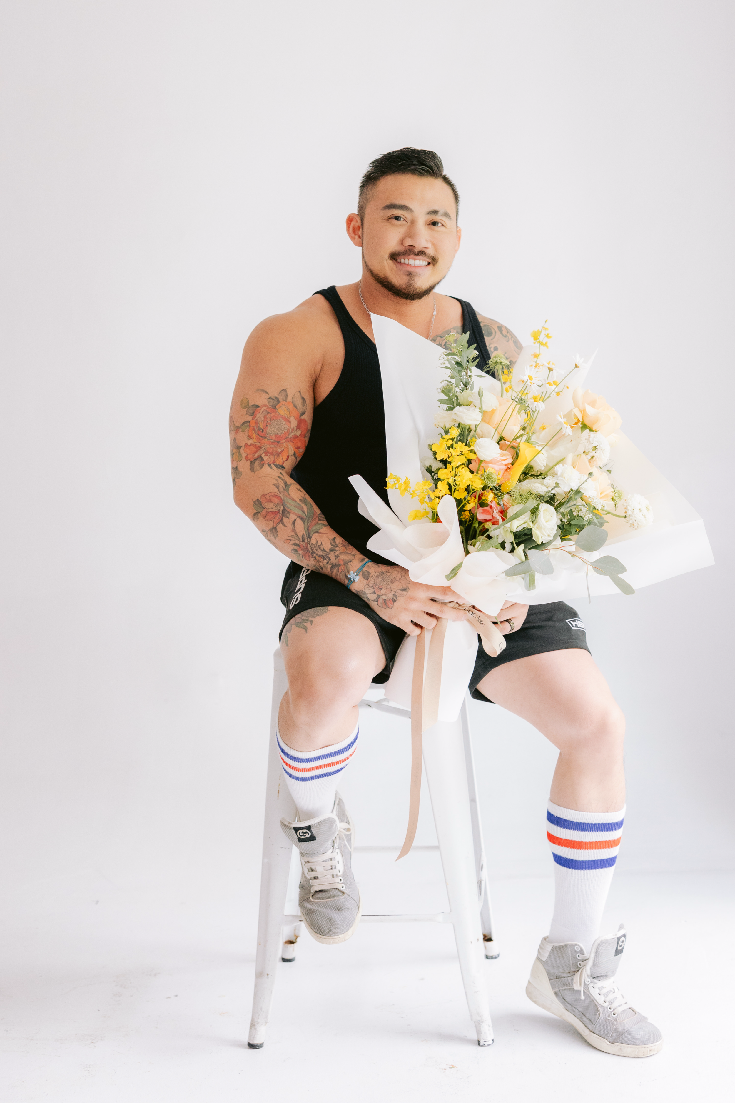 A smiling man sitting on a white chair, holding a large bouquet of flowers, wearing a black tank top, black shorts, white athletic shoes, and knee-high socks with red and blue stripes, set against a plain white background.
