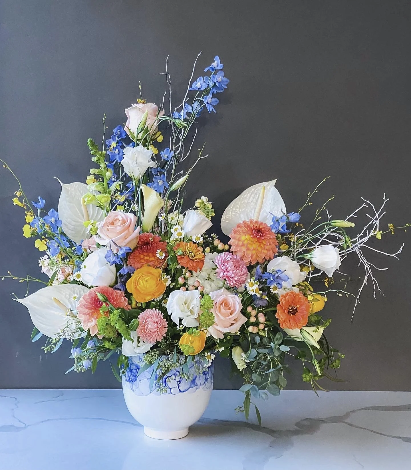 A large colorful flower arrangement in a white ceramic vase featuring pink roses, white calla lilies, orange and pink dahlias, blue delphiniums, and various greenery, set against a dark gray background.